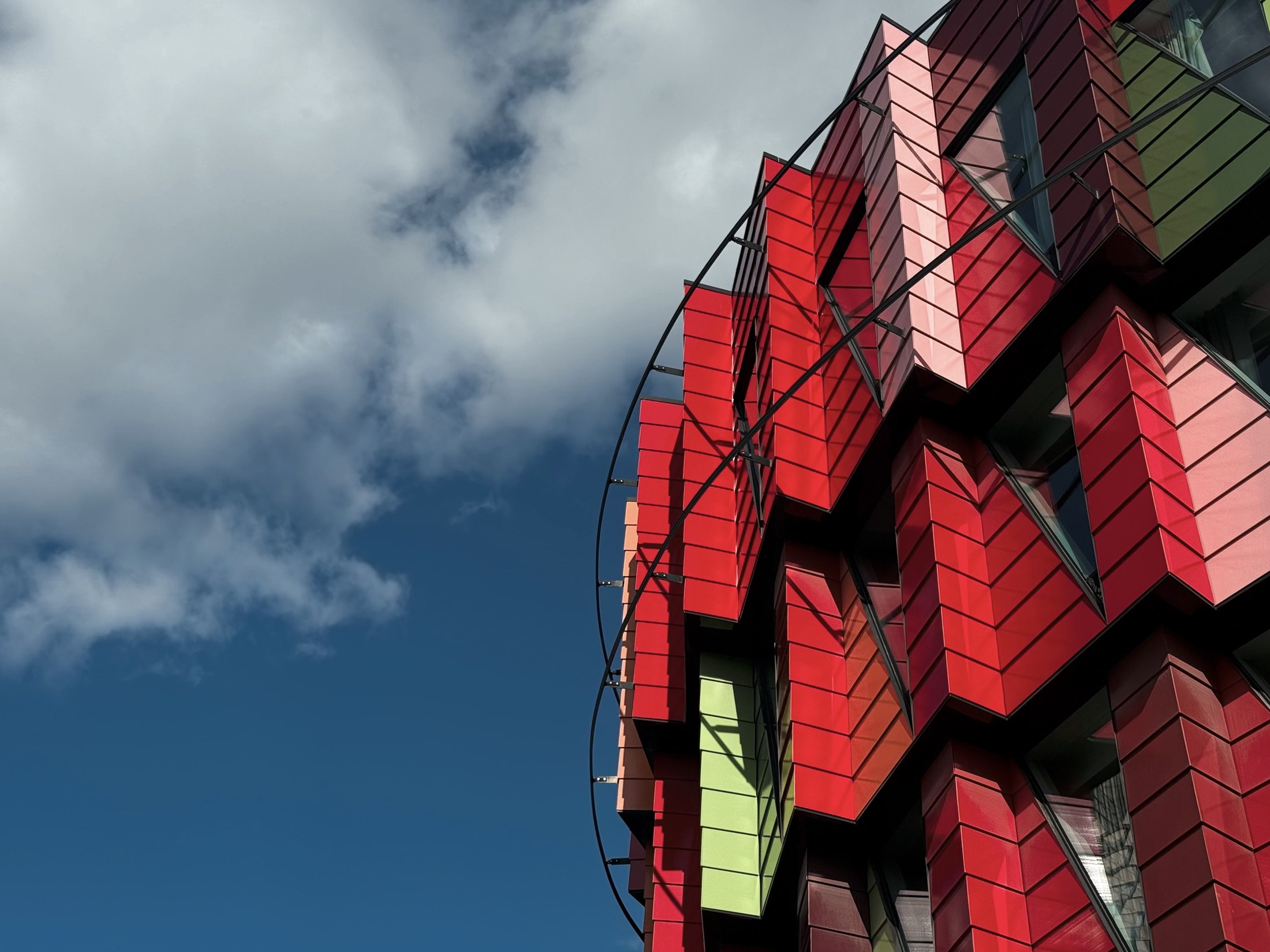 The Kuggen building at Lindholmen in Gothenburg, a rounded facade of faceted panels in reds, pinks and green, photographed from below against a blue sky where a large white cloud fills the left half of the frame.