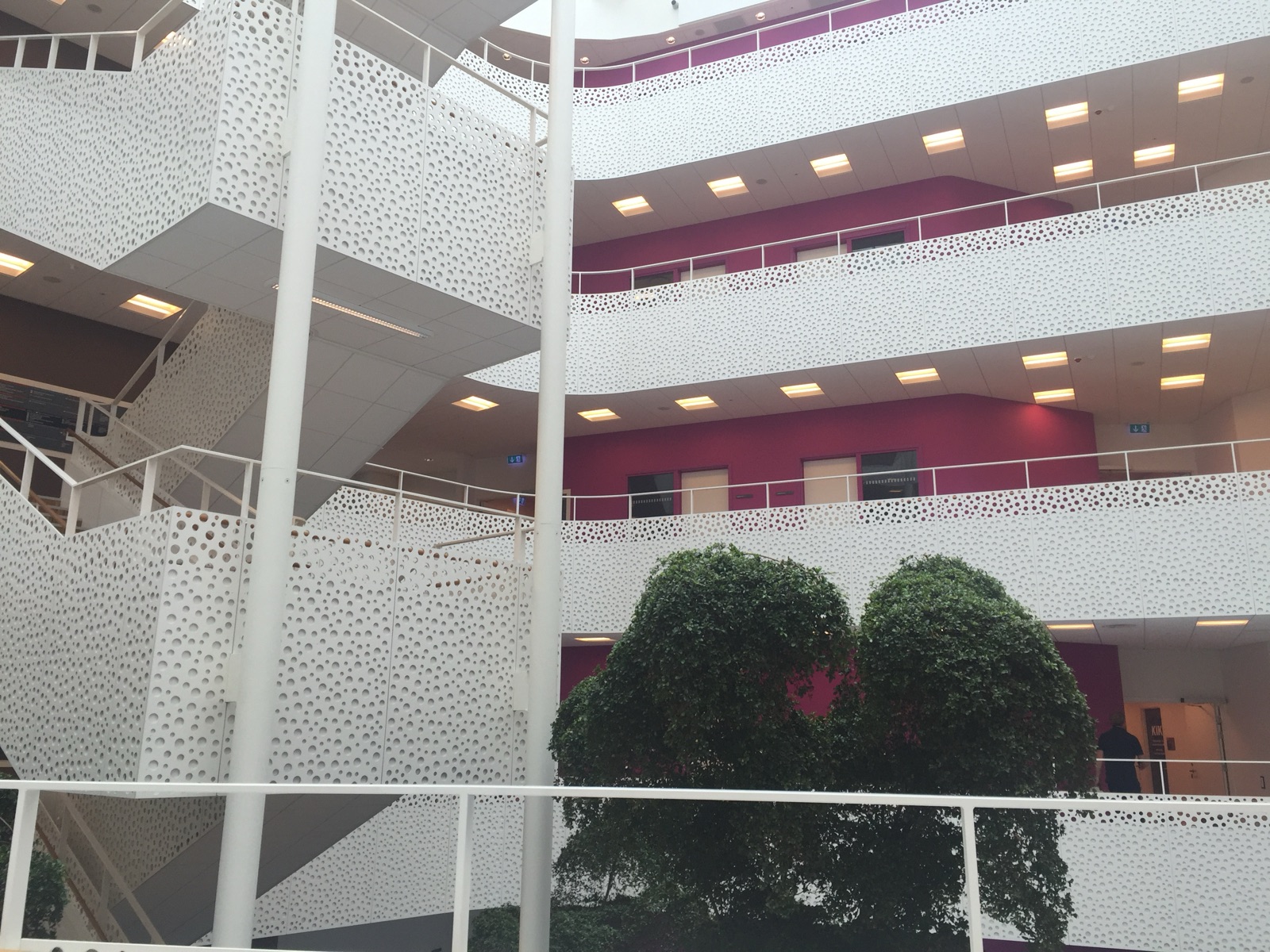 A level of the atrium in Handicaporganisationernes Hus, layered white perforated balconies against deep pink accent walls, with indoor trees below.