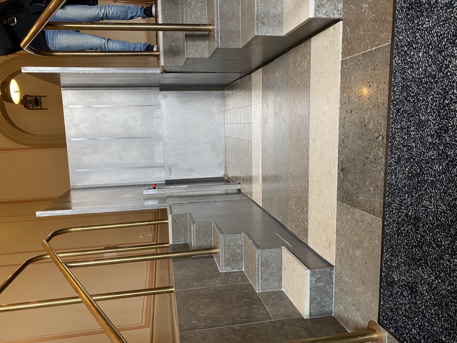 Flexstep stair lift in a protected historic building, a flat steel platform recessed into the marble floor with folded stair treads along one edge and brass railings.
