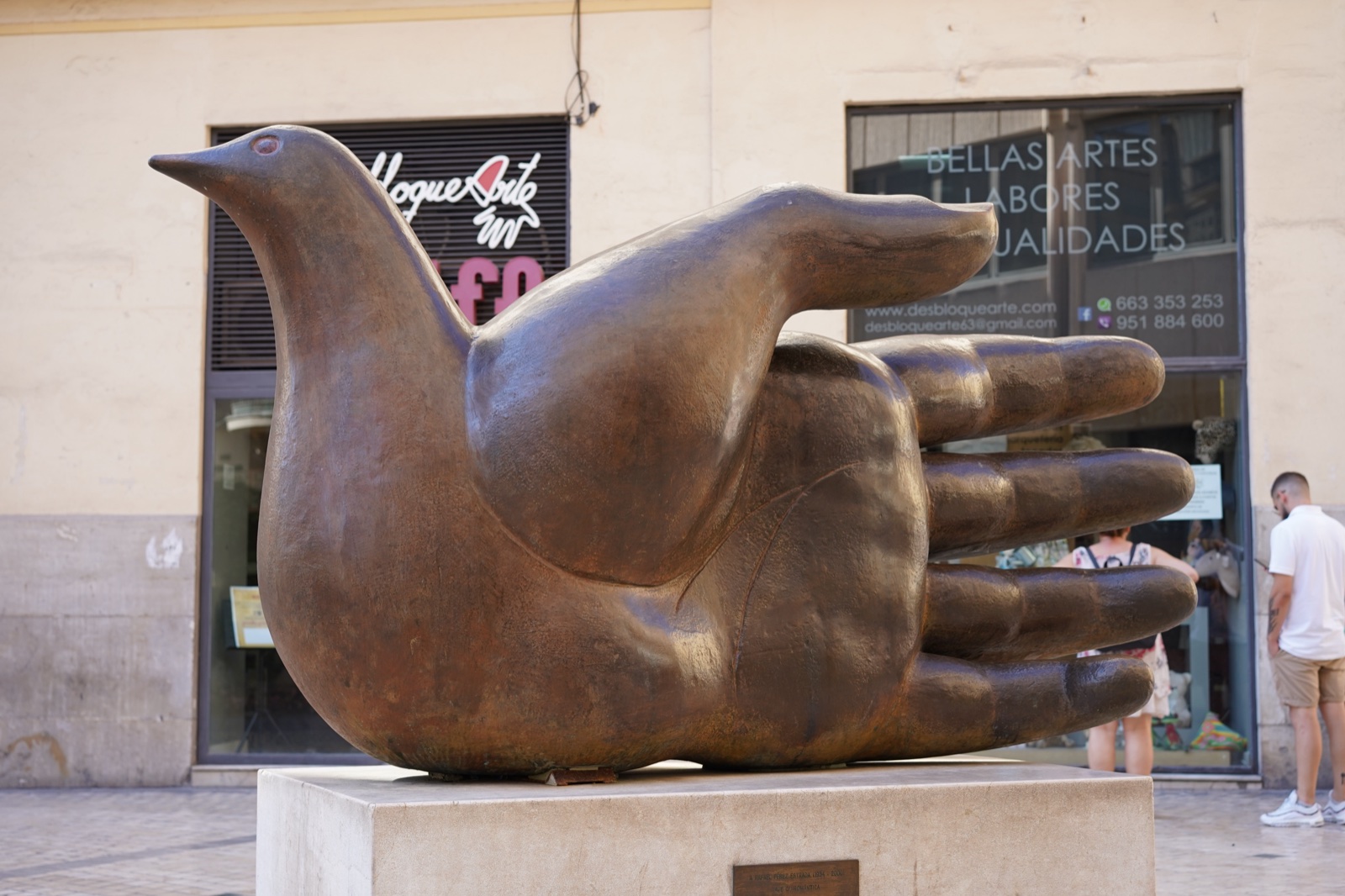 Bronze sculpture of a bird and an open hand fused into one body, on a low plinth in a pedestrian street