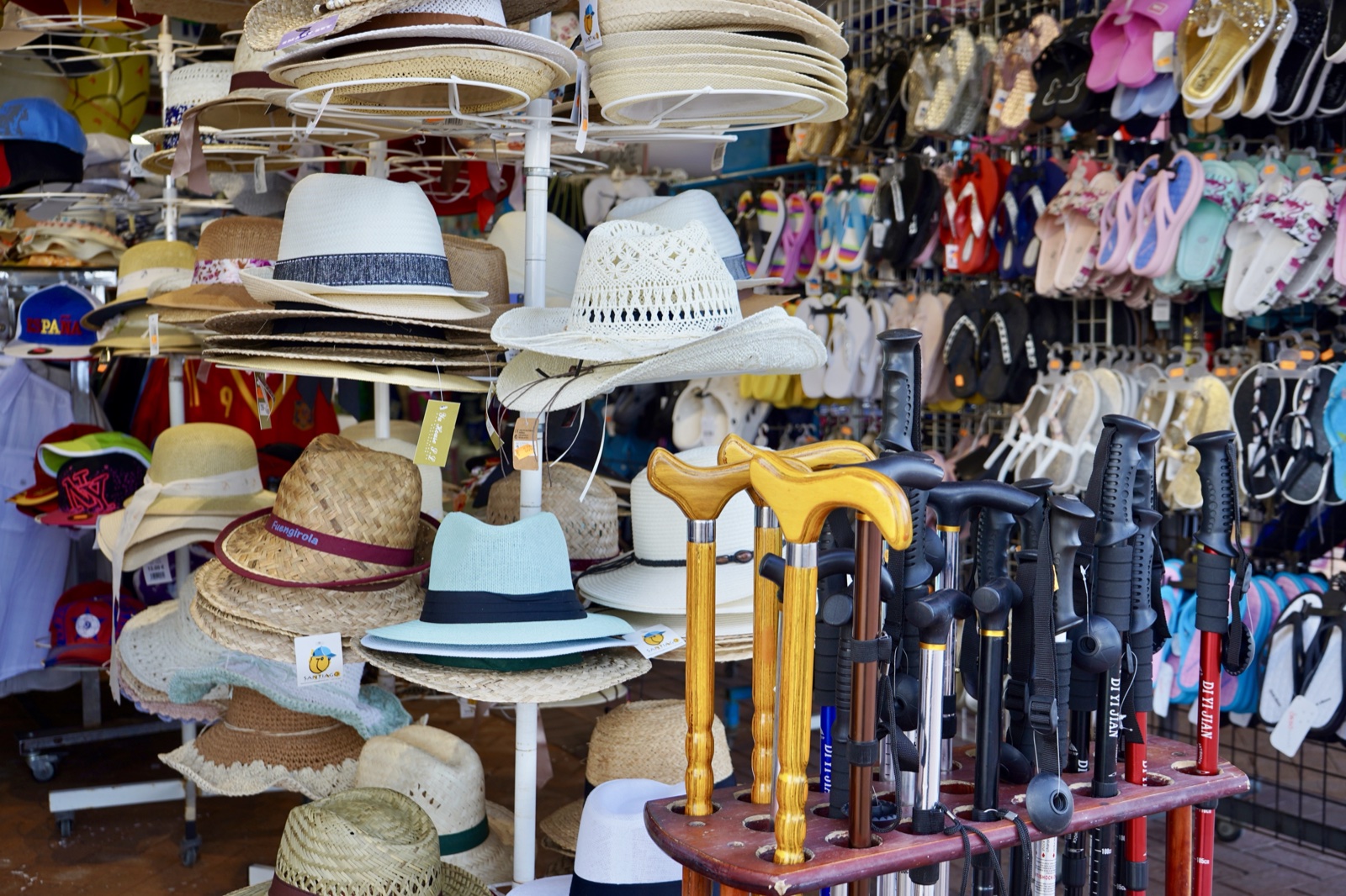 Street market stall in Fuengirola, straw hats on rotating racks and walls of flip-flops. In front, a wooden stand where wooden walking canes stand interleaved with black trekking poles.