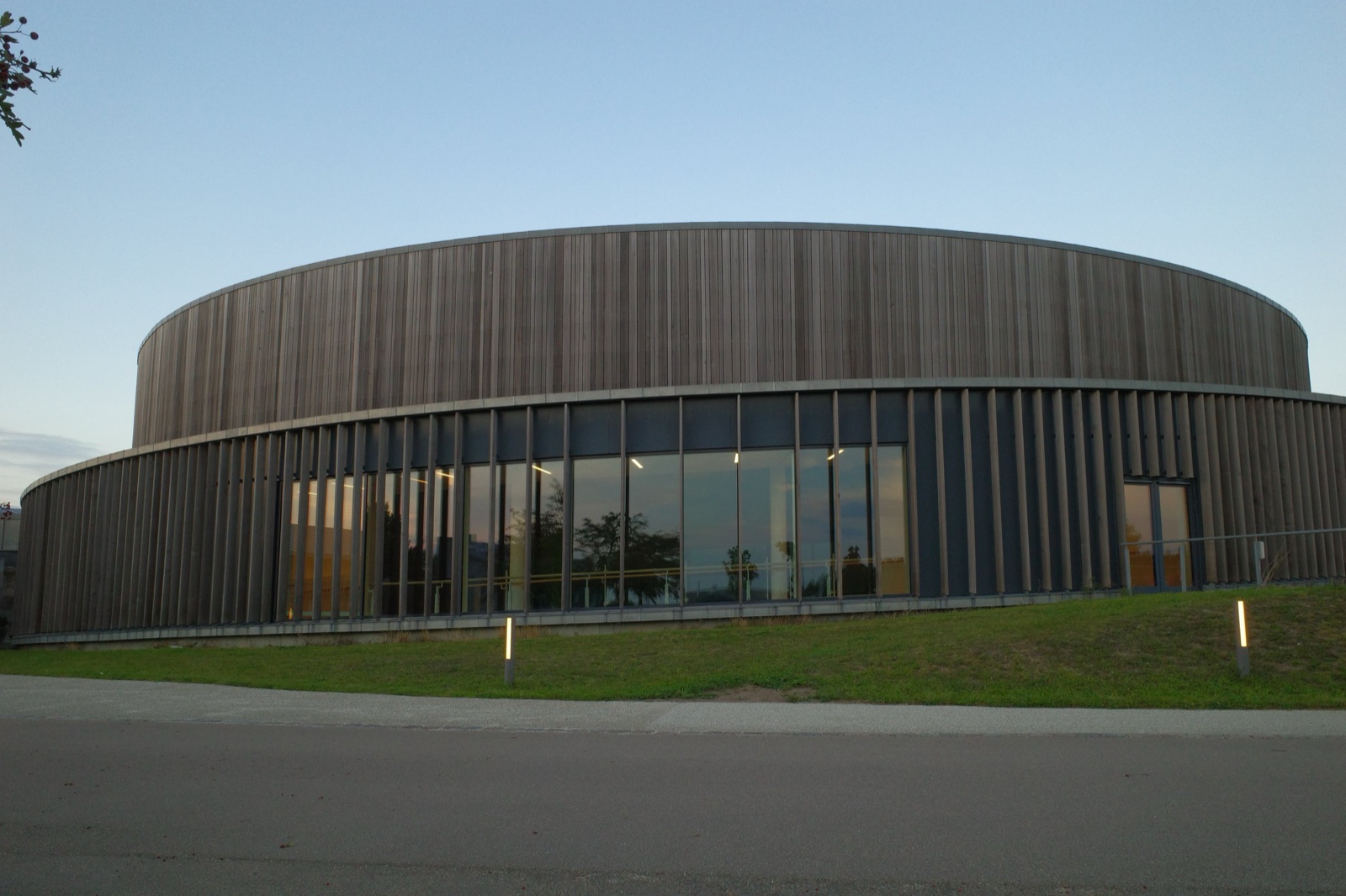 Circular timber-clad sports hall at dusk with a glazed ground floor and a handrail visible inside the glass
