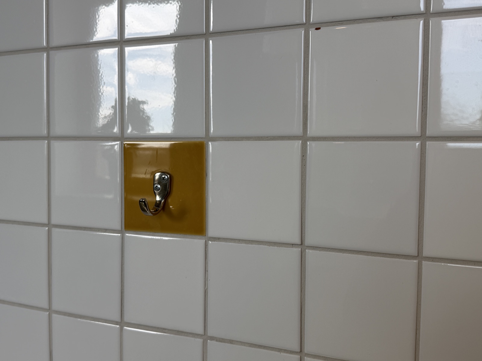 White tiled wall with a single ochre tile carrying a chrome hook at seated height