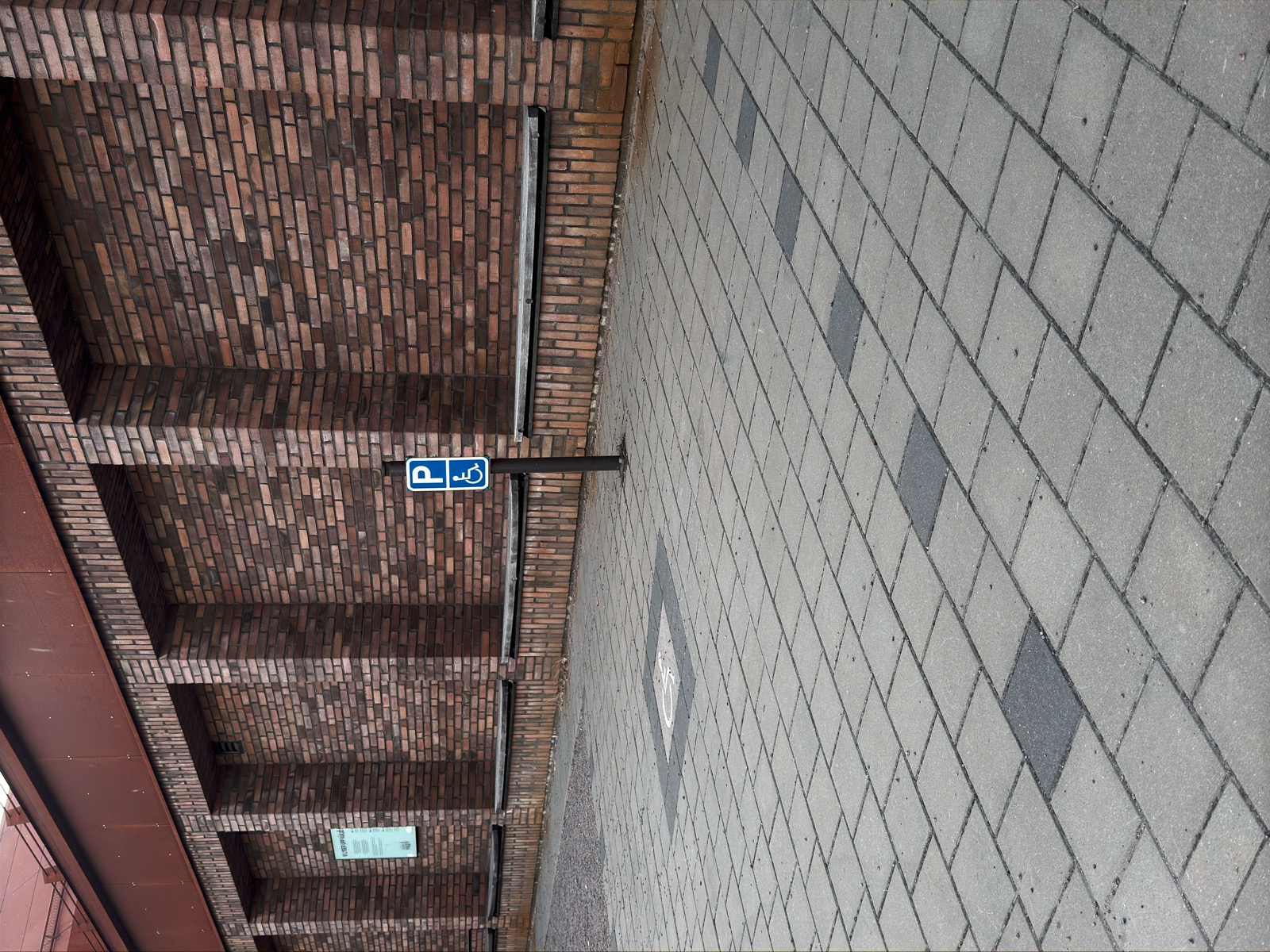 An accessible parking bay beside a brick building: a P sign with the wheelchair symbol on a post, and the same symbol inlaid in the paving of the bay