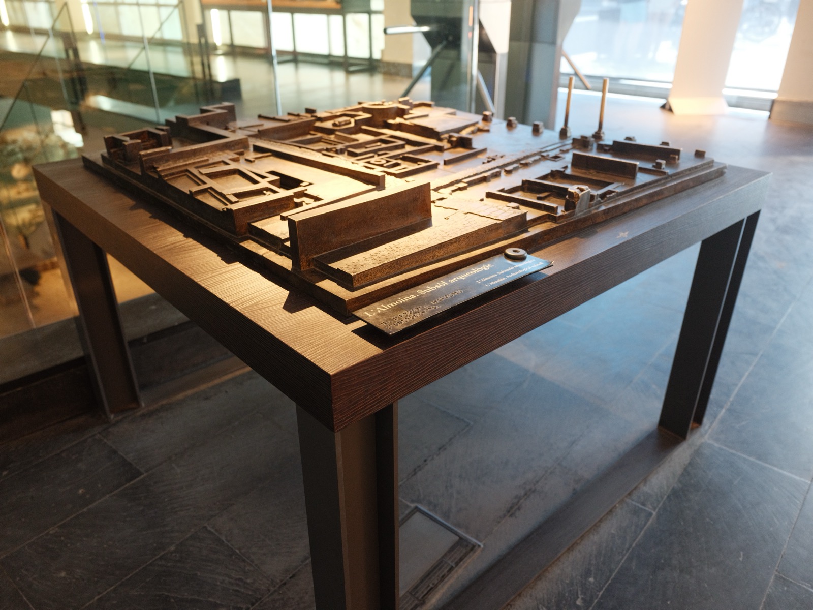 Bronze tactile model of the L’Almoina archaeological site on an open table, with the site name in raised letters and braille on the label.