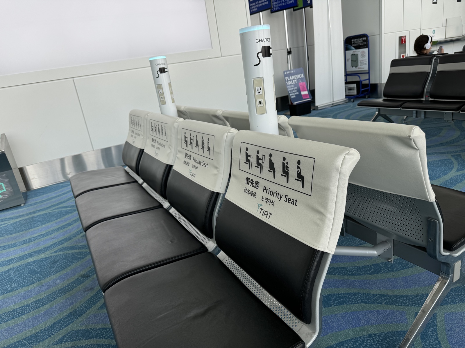 Gate seating at Haneda airport. In a row of identical black bench seats, four seats wear white slip-on covers printed Priority Seat in four languages, with five seated pictograms, a person with a cane, one with a crutch, a pregnant woman, one holding an infant, and one with a heart-and-cross mark. Charging pillars stand between the seats.