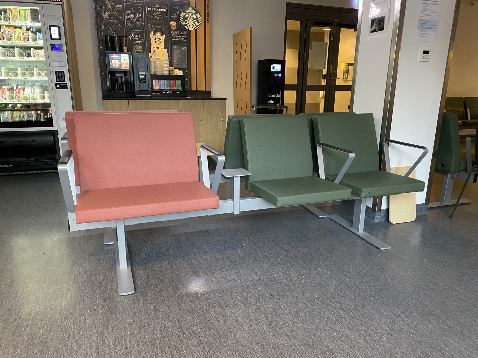 Beam seating in a Swedish hospital waiting area. On one shared steel beam, a double-width seat in dusty pink and two single seats in green with armrests, joined by a small table plate. A coffee corner and vending machine behind.