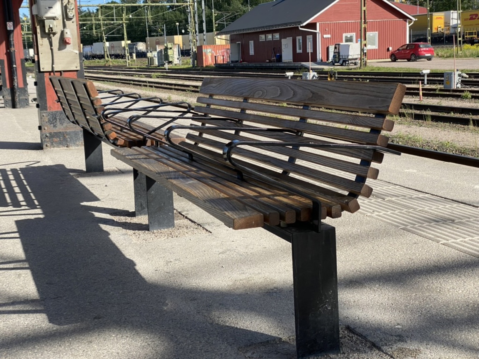 Two wooden-slatted benches on a Swedish rail platform, each with a curved ergonomic profile and a continuous black steel tube that weaves along the bench and rises above seat level partway along, dividing the seat.