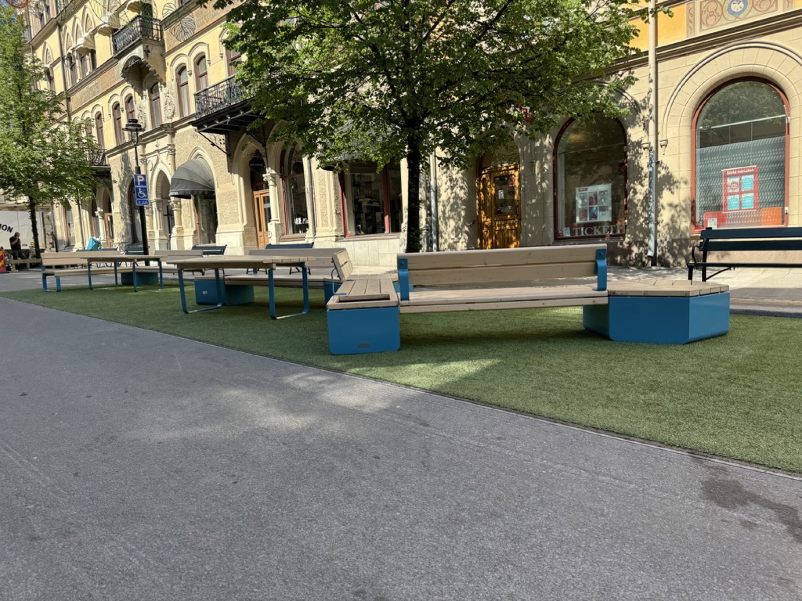 Blue-framed benches, tables and bench-boxes on artificial grass forming a parklet in a Swedish pedestrian street, historic facades behind. An accessible-parking sign stands at the edge of the turf.