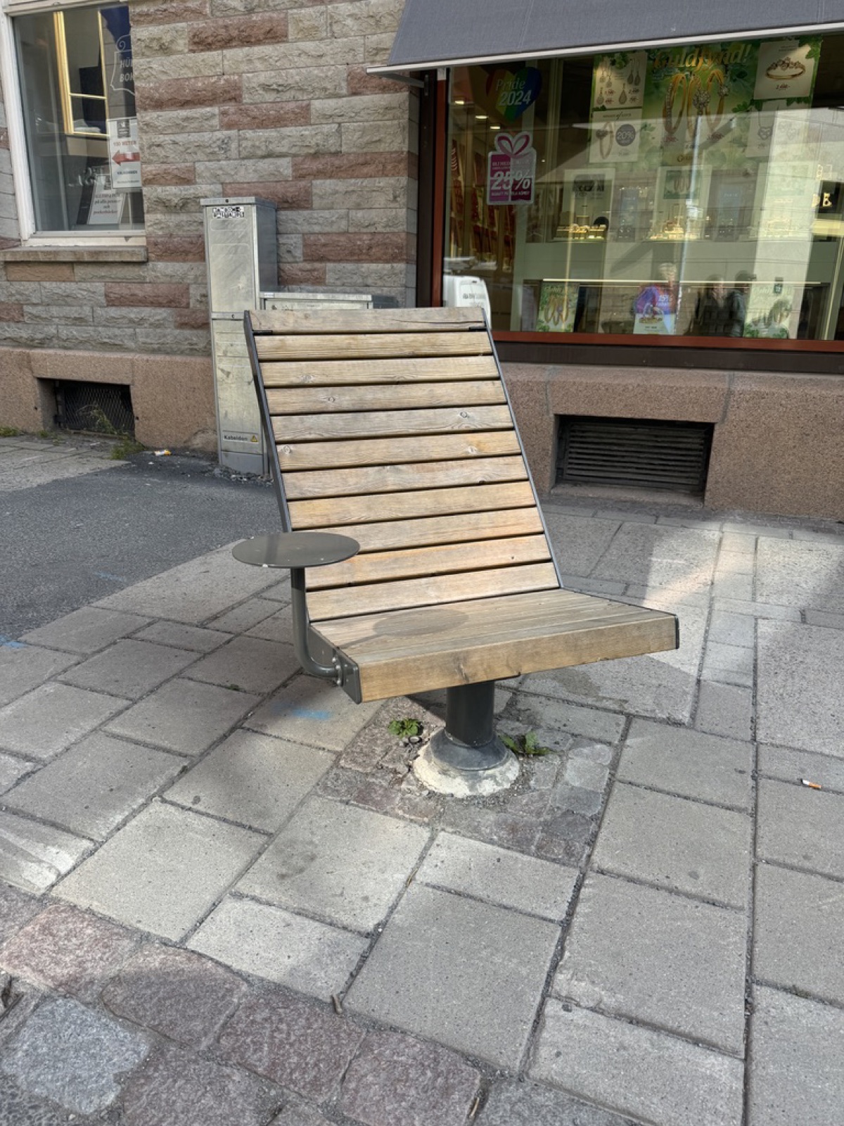 Single-seat wooden bench on a centre pedestal on a Swedish sidewalk, with a high slatted back and a small round metal side table attached at the sitter's right.
