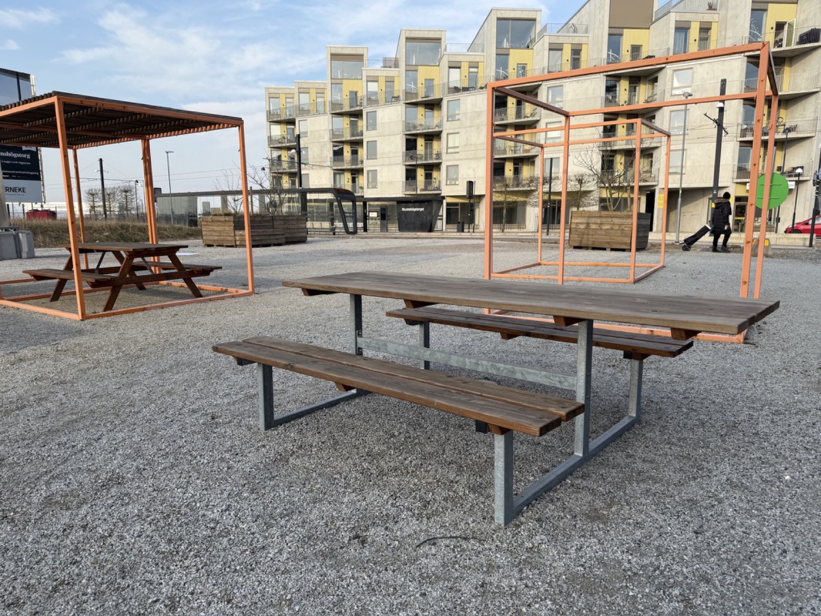Picnic table with a galvanised steel frame on a gravel square between apartment blocks. The tabletop extends beyond the benches at the near end, leaving a free open edge. Orange steel-cube pergolas behind.