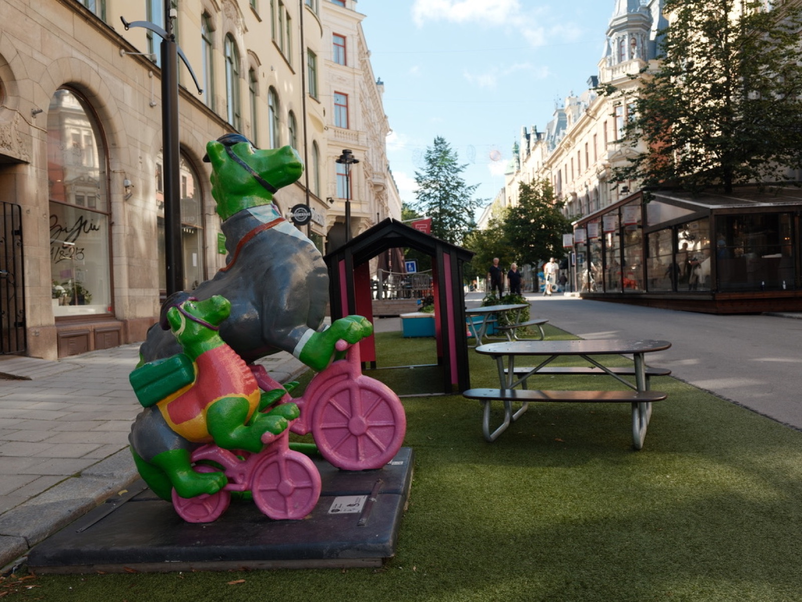 Summer pedestrian street in Sundsvall with artificial turf, picnic tables, a small wooden playhouse, and two dragon sculptures riding pink bicycles. Cafes and stone-town facades along the street.