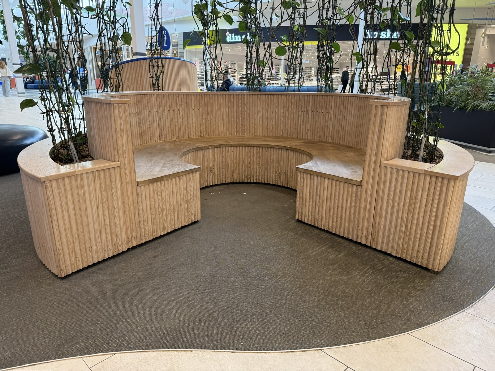 Semi-circular wooden bench in a shopping mall, built from light vertical slats with integrated planters and an open front.