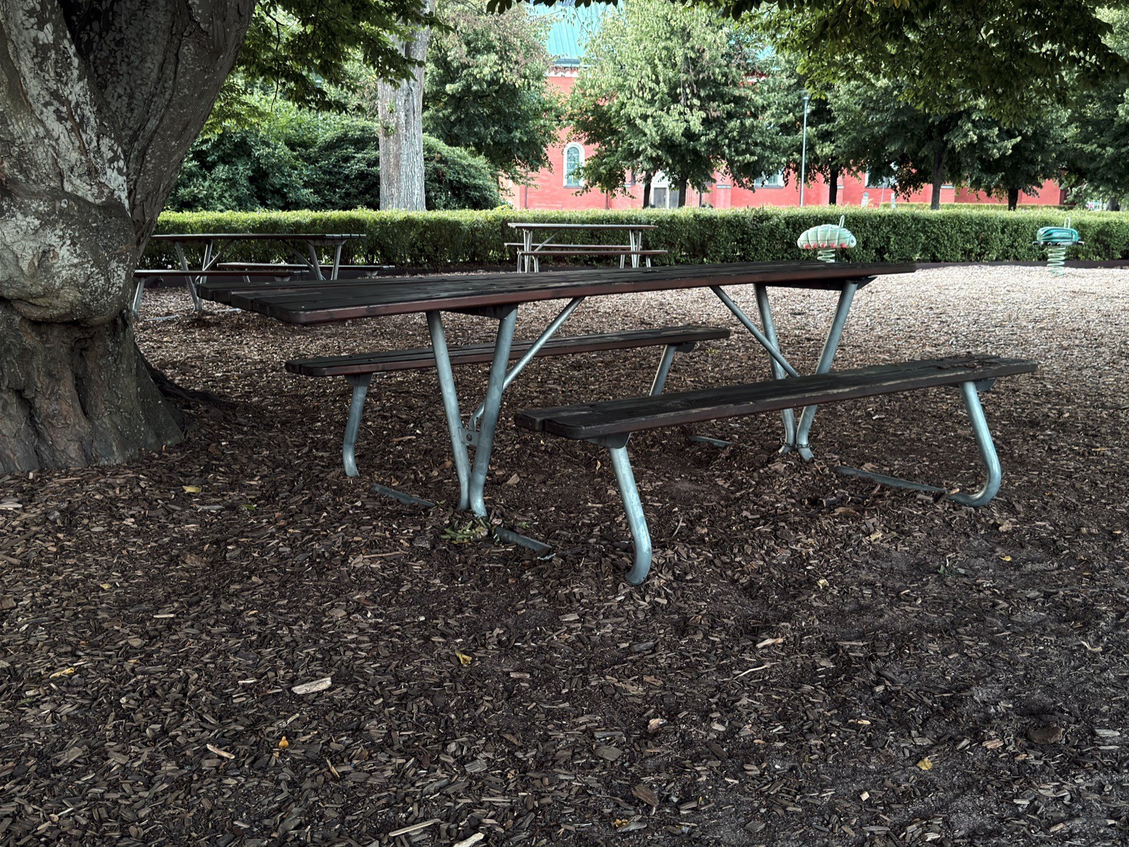Wooden picnic table on a metal frame under a large tree in a Växjö park. The tabletop extends well past the benches at the near end, leaving an open place with no bench in the way.