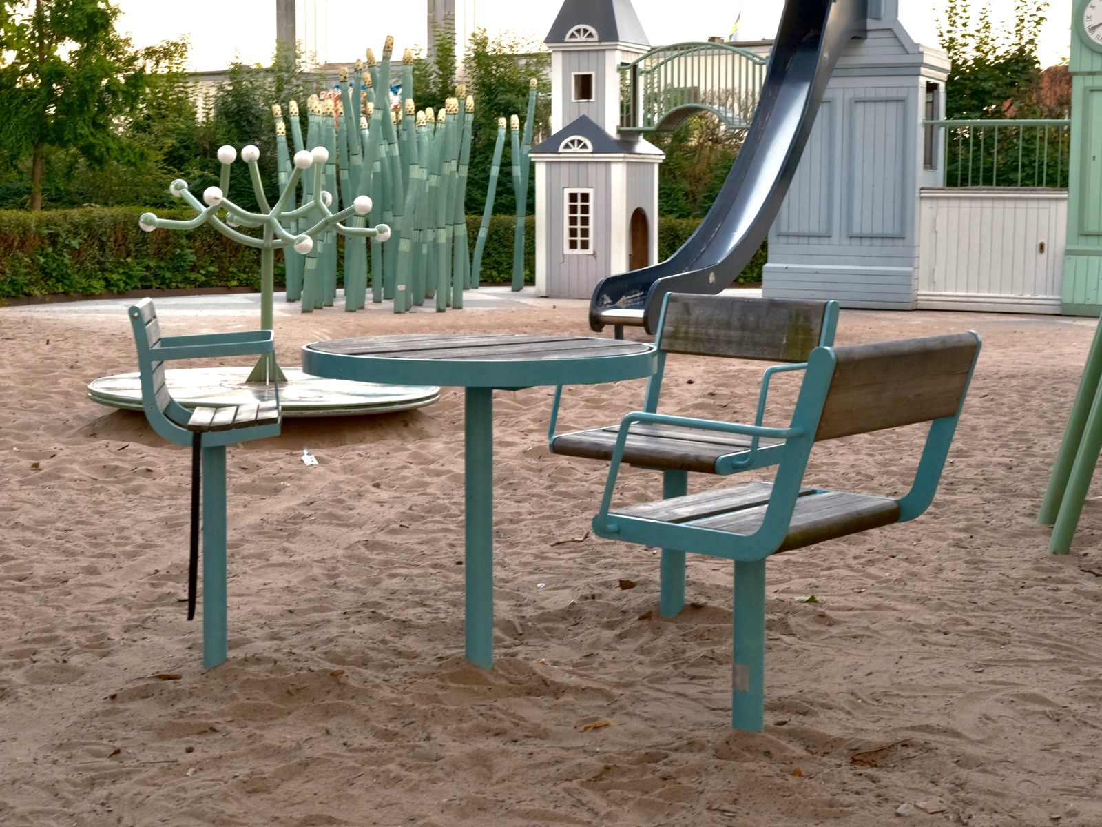 Round table on a single centre pedestal in the sand of a playground in Växjö, with three chairs around it, one of them a raised baby chair, and a fourth place left open. Play houses and a slide behind.