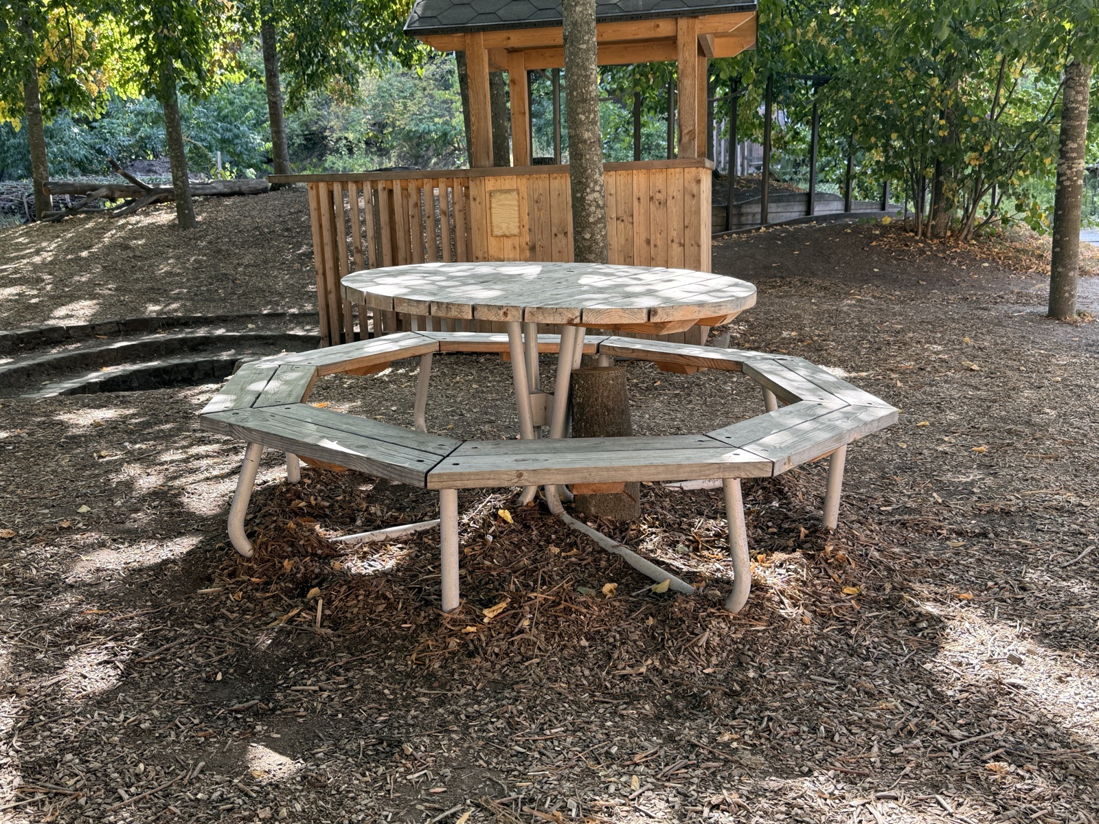 Round wooden table ringed by a continuous octagonal bench on bark mulch under a tree. The bench runs the whole way round with no gap.