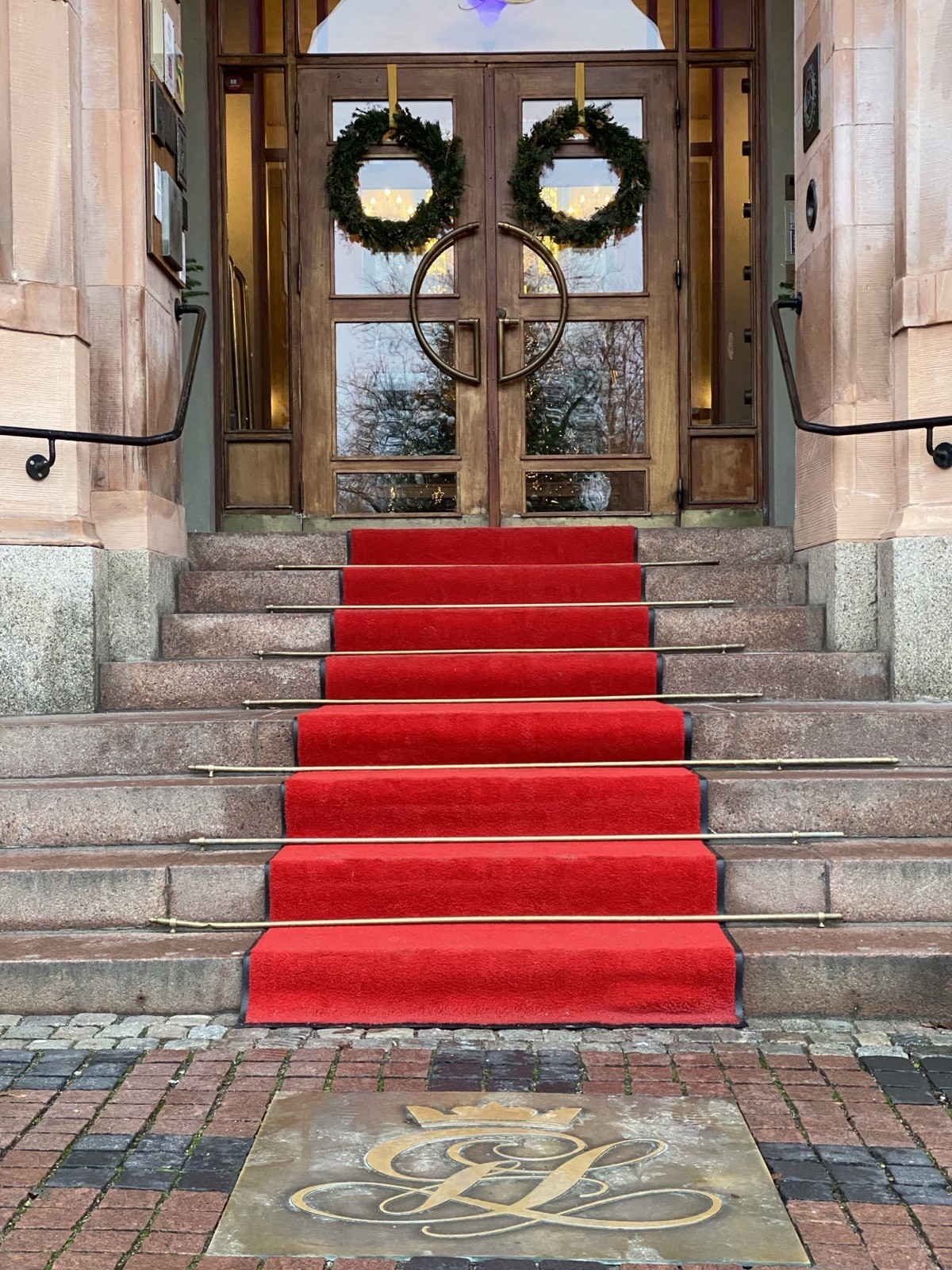 Grand hotel entrance with an ornate arched stone portal. A red carpet runner goes up the stone stairs to double doors with Christmas wreaths.