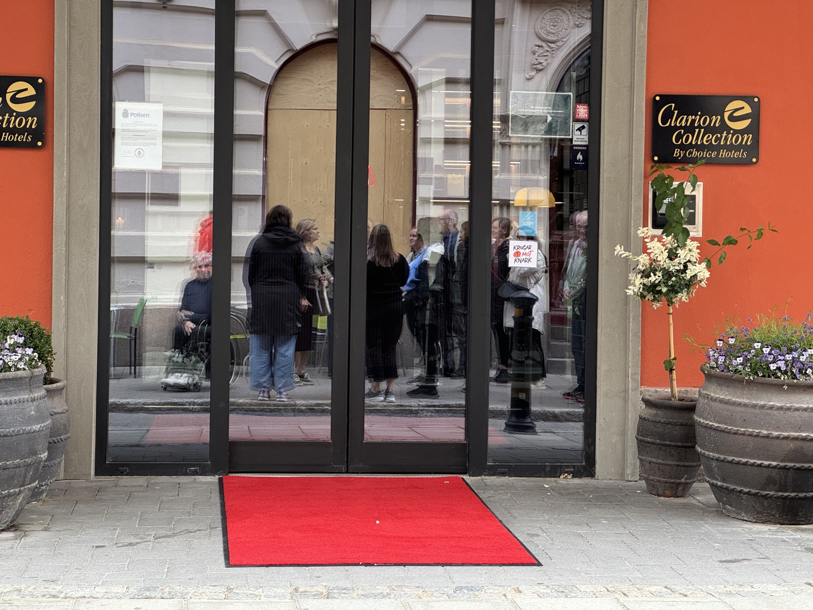 Hotel entrance in summer. A red carpet mat on flat pavement at street level, summer flowers in pots, people reflected in the glass doors.