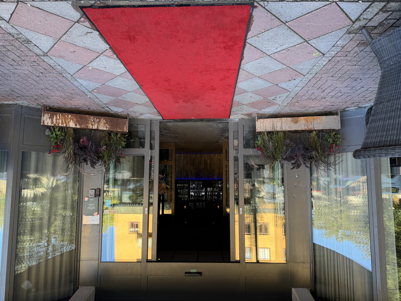 Entrance on a walking street in evening light. A red carpet on flat stone paving leads to wide-open glass doors flanked by flower arrangements.