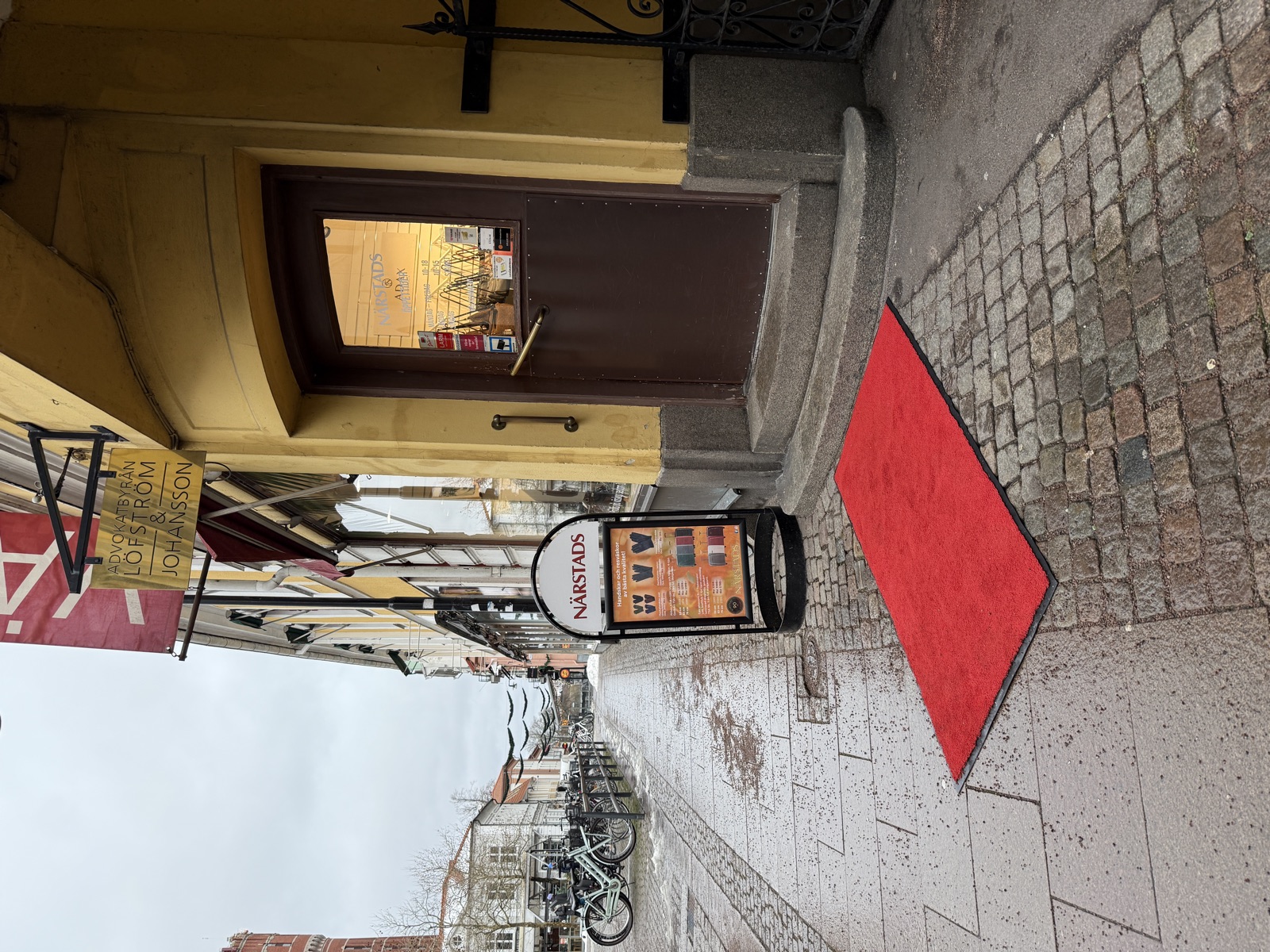 Small-town cafe entrance. A red carpet mat on the cobblestone pavement in front of stone steps leading up to the doorway.