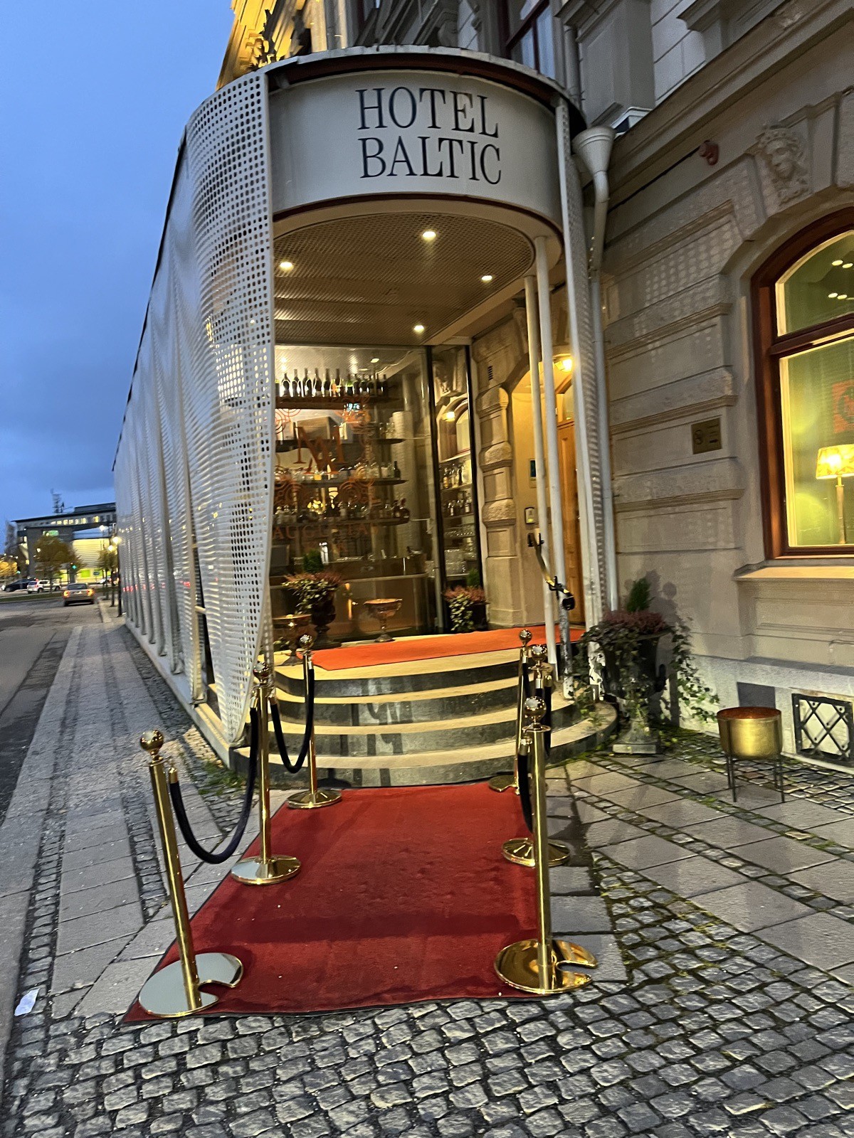 Hotel entrance in the evening, a curved white canopy reading Hotel Baltic over an ornate stone facade. A red carpet lies on the flat cobblestones inside a frame of brass stanchions with ropes, and continues on the landing by the doors. The stone steps between them are bare.