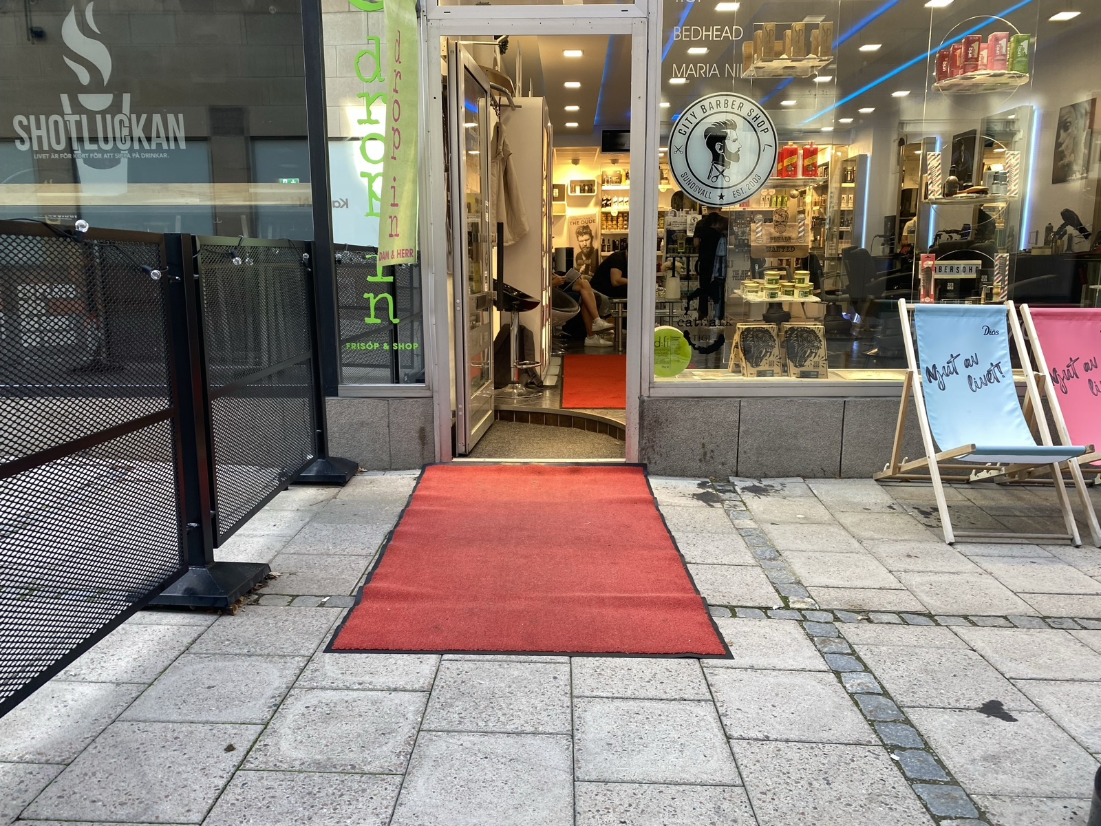 Barber shop entrance on a pedestrian street. A red carpet mat lies flat on the pavement in front of the open door. Inside the narrow doorway there is a step up into the shop, where a second red mat continues.