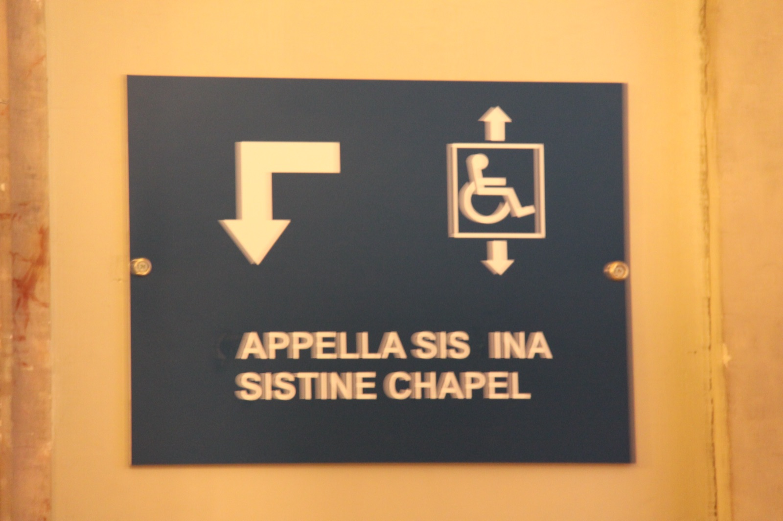 Wayfinding sign at the Vatican Museums: CAPPELLA SISTINA, SISTINE CHAPEL in relief letters, several of them fallen off, with a turn-then-down arrow for the stairs and a wheelchair-in-elevator icon with up and down arrows. Dark sign on a marble wall.