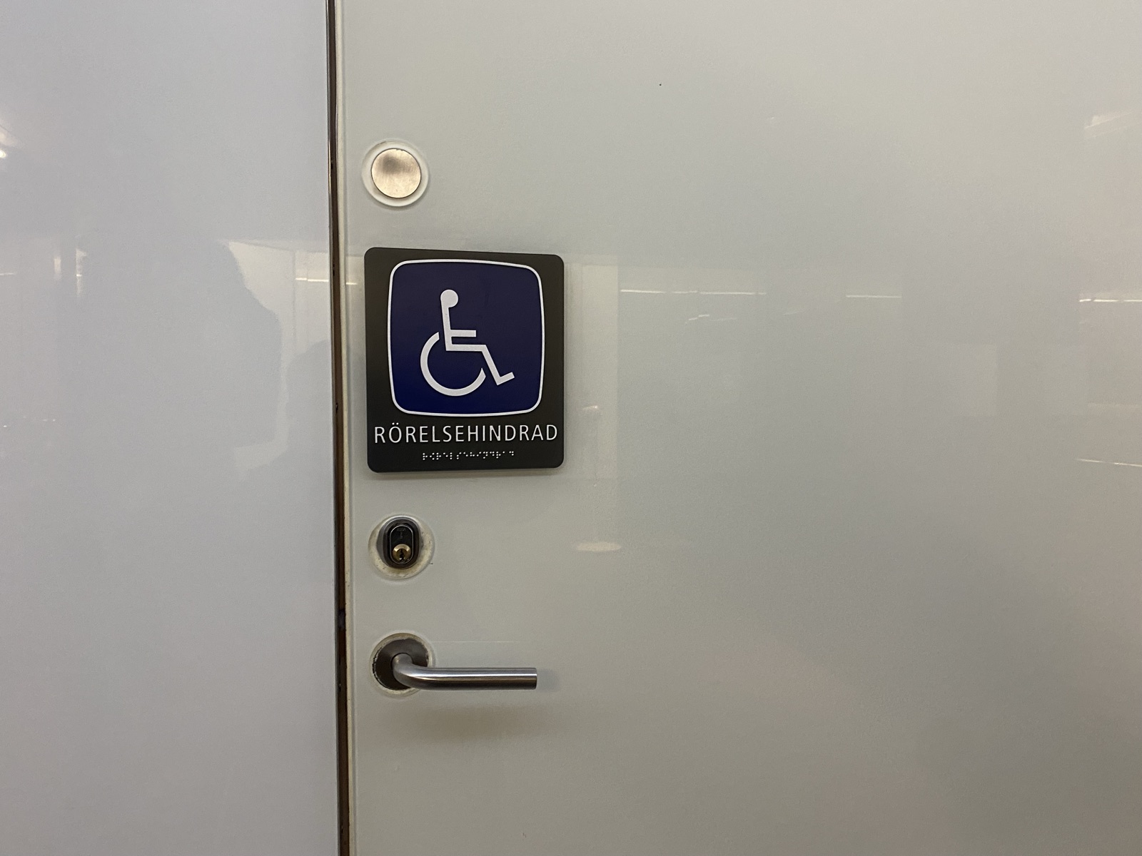 White door with a blue/black sign: wheelchair symbol above the word "RÖRELSEHINDRAD" (mobility impaired) with braille below. Metal handle and lock.