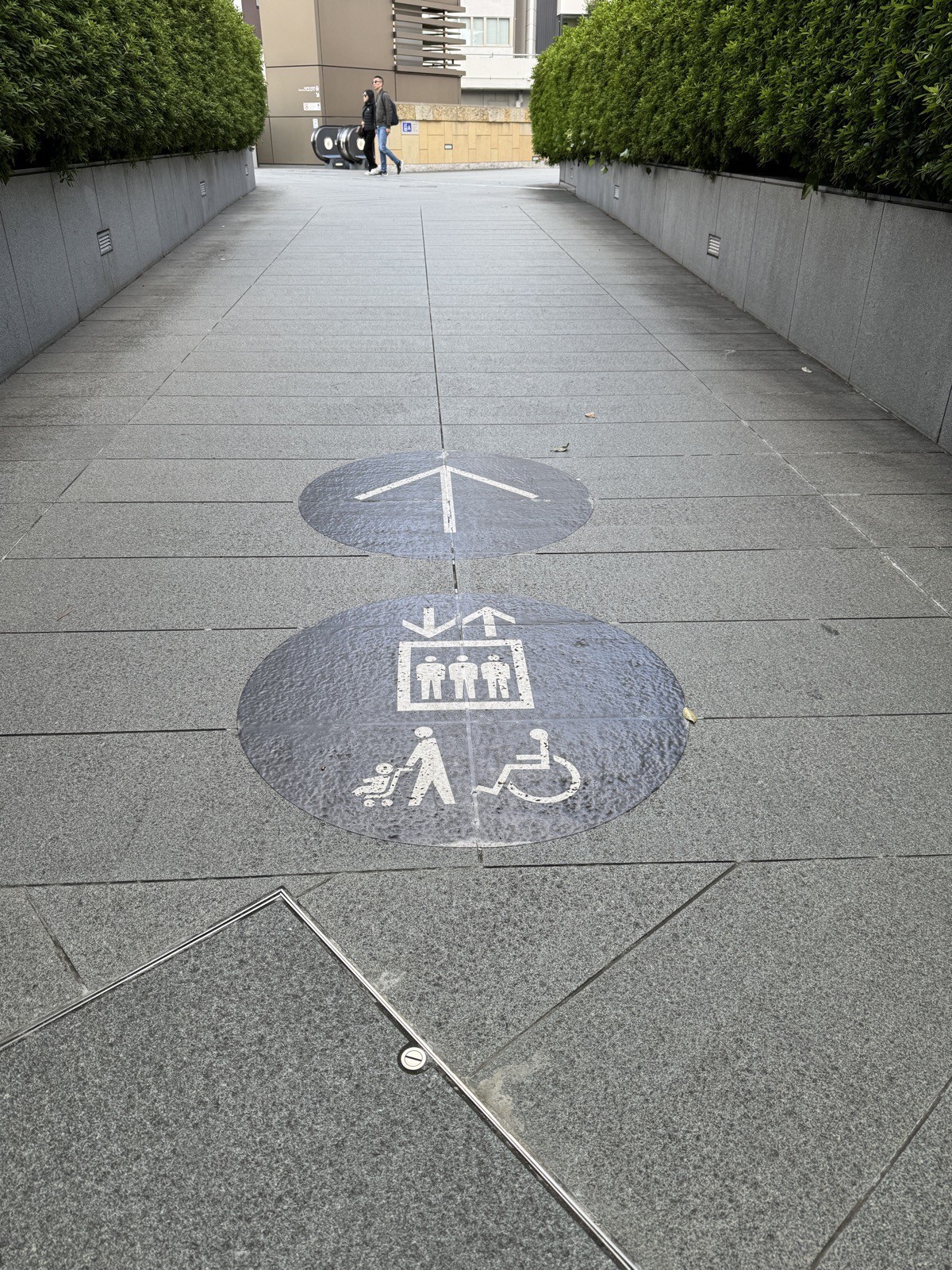 Two circles painted on grey stone pavement in Japan, one with a directional arrow, one with an elevator icon, a person pushing a pram, and a wheelchair user.