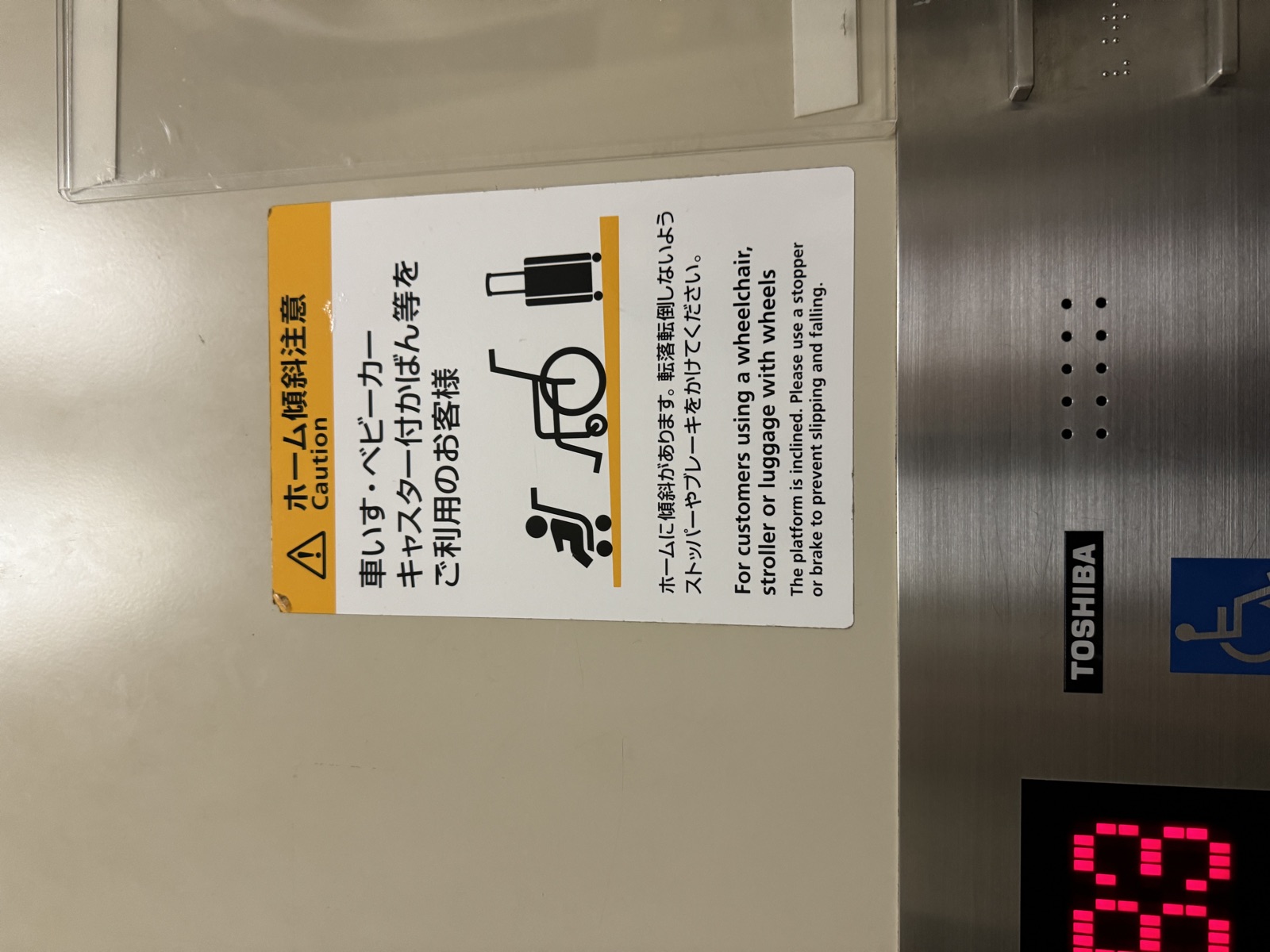 Yellow and white caution sticker inside a Japanese station elevator, with a stroller, an empty wheelchair, and a wheeled suitcase standing on a sloping line, warning that the platform is inclined.