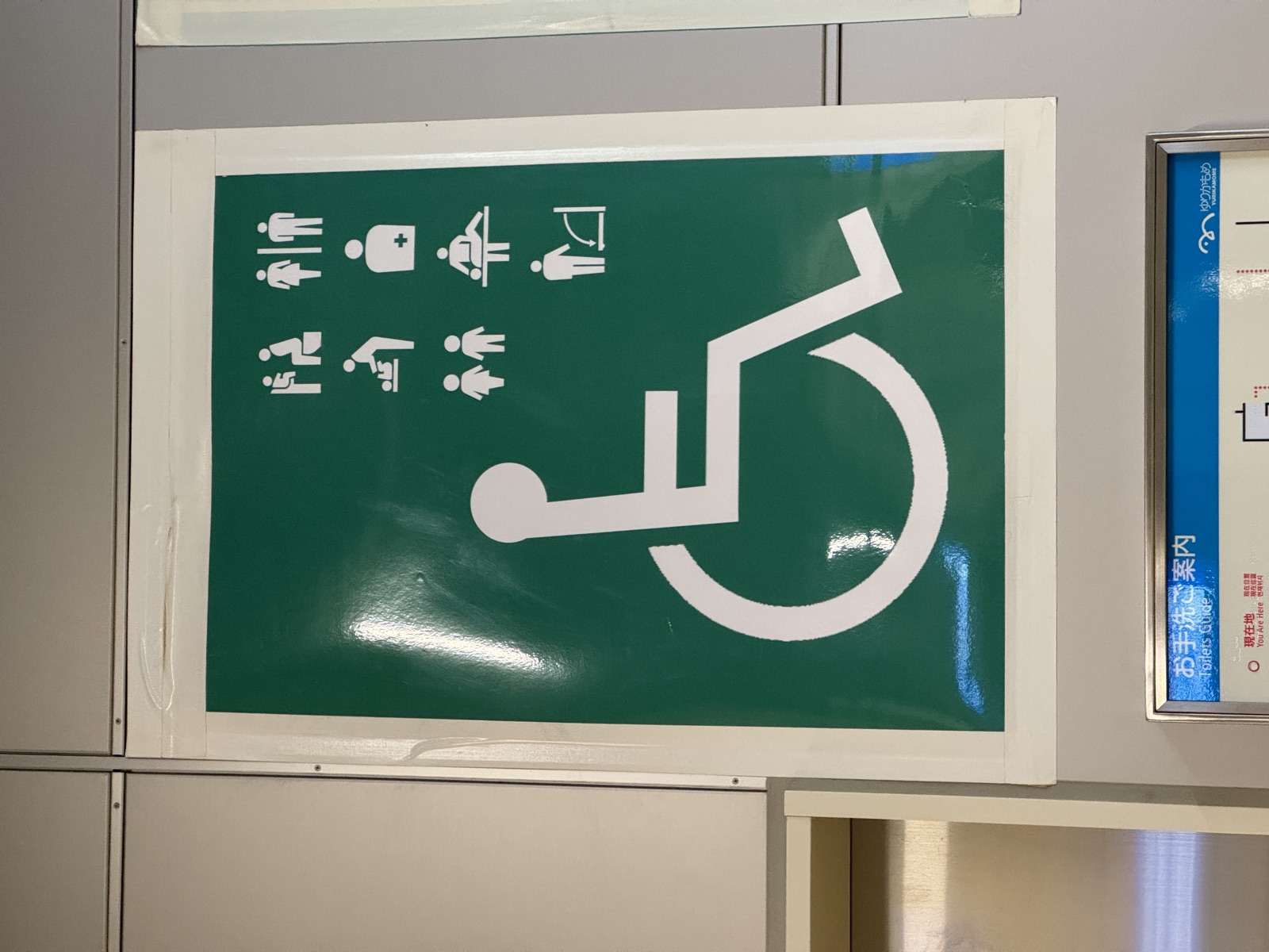 Green sign in a Japanese station, a large white wheelchair symbol with seven smaller pictograms in rows beside it, toilet users, a women-and-men pair, baby changing, a torso with a medical cross, two children, an adult changing bench, and a fold-down fixture.