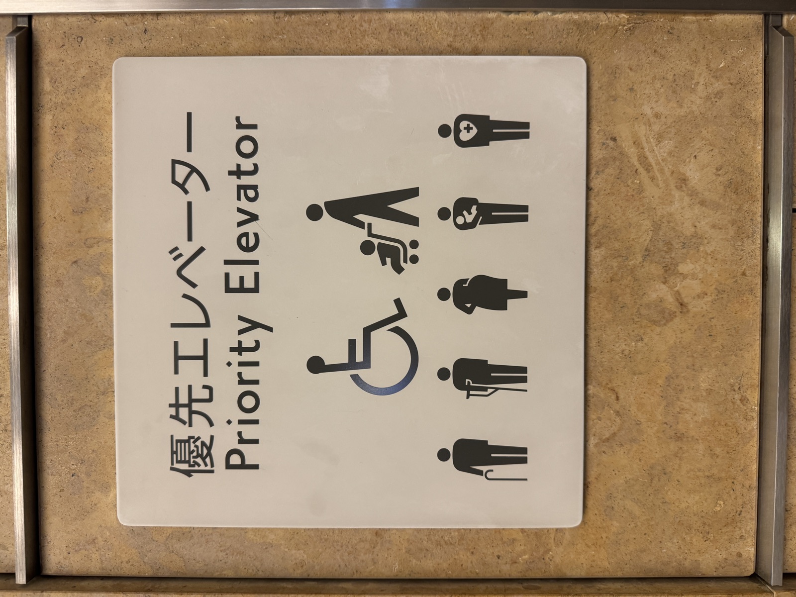 Japanese Priority Elevator plate on a beige stone wall. A large wheelchair symbol and a parent pushing a stroller on top. Below them five smaller figures, one with a cane, one with a crutch, one pregnant, one carrying a baby, and one with a heart-and-cross mark for non-visible conditions.