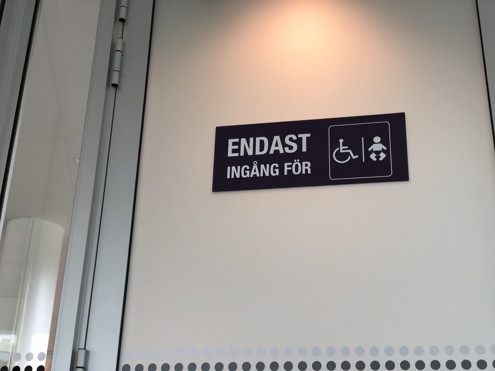 Dark purple plate by a Swedish glass entrance reading ENDAST INGÅNG FÖR, Only entrance for, beside a rounded box holding a wheelchair symbol and an infant figure separated by a line. Rows of contrast dots run across the glass below.