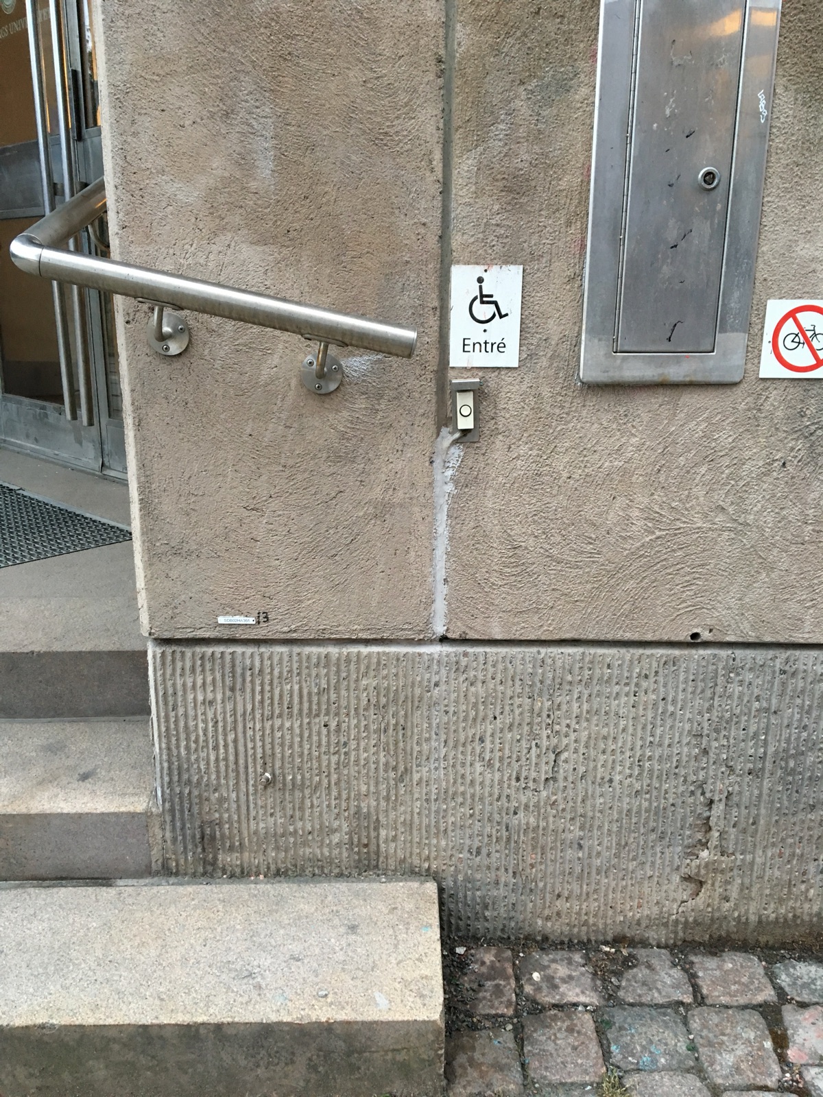 Stone entrance steps at a Swedish university building. Beside the steps, a small sign with a wheelchair symbol and the Swedish word Entré, with a call button beneath it. Metal handrail over weathered concrete.