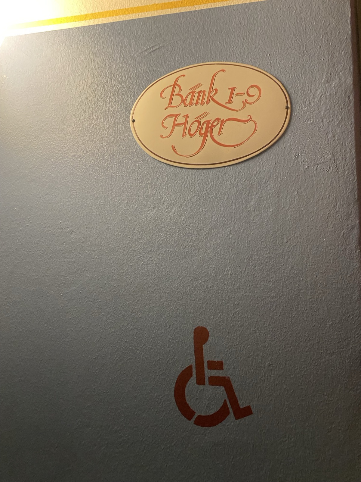 Church interior detail: on a grey-blue wall, a cream oval plaque with Bänk 1–9 Höger, Pews 1–9 Right, in red calligraphic script, and below it a wheelchair symbol stencilled directly onto the wall in dark red paint, matched to the church's palette.