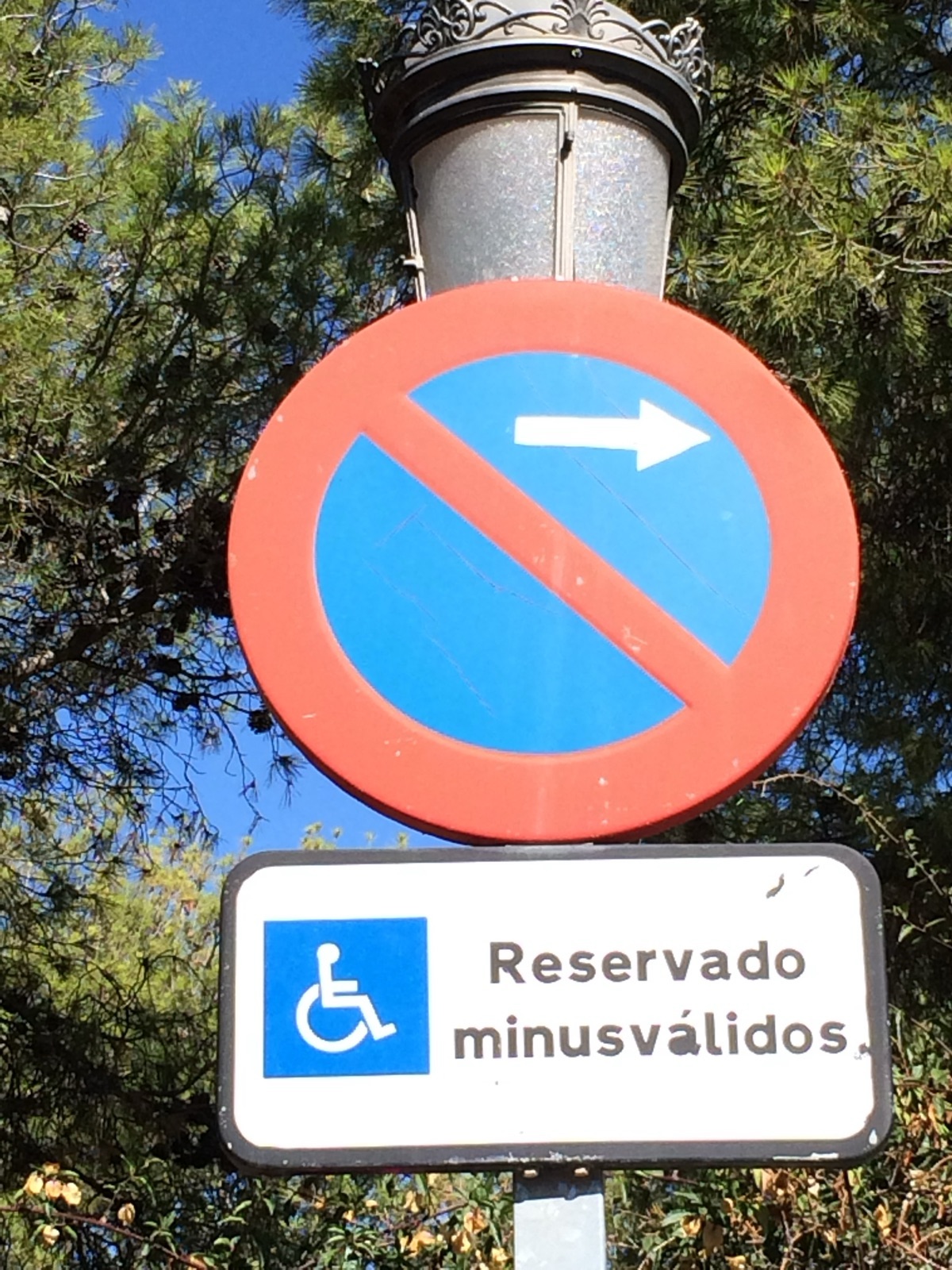 Spanish street sign on an ornate lamp post: a no-parking circle with an arrow, and beneath it a white plate reading Reservado minusválidos, Reserved for the disabled, with a blue wheelchair square.