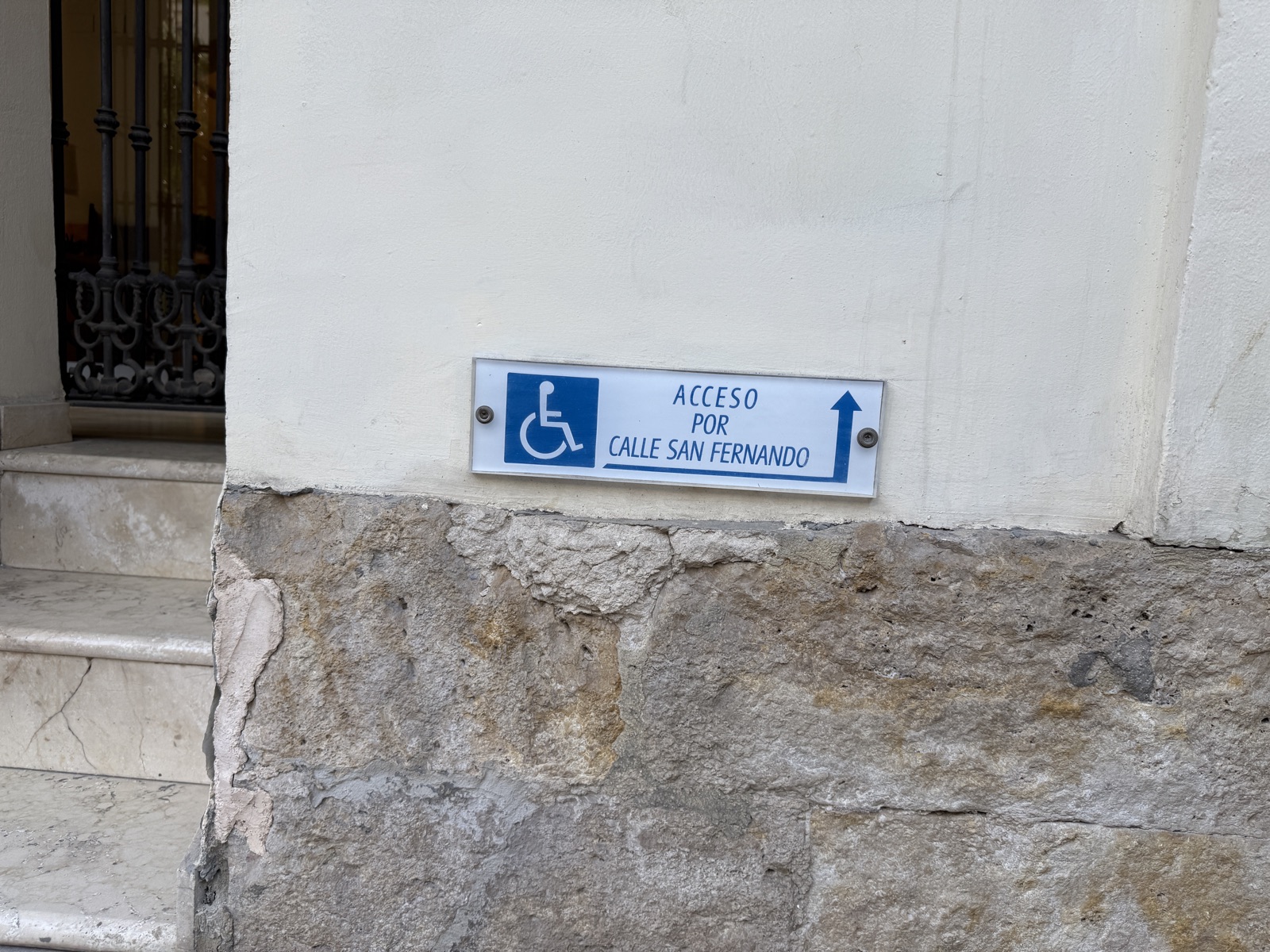 Sign on a stone wall in Spain, a wheelchair symbol and the text Acceso por Calle San Fernando with an arrow. Steps are visible beside the sign.