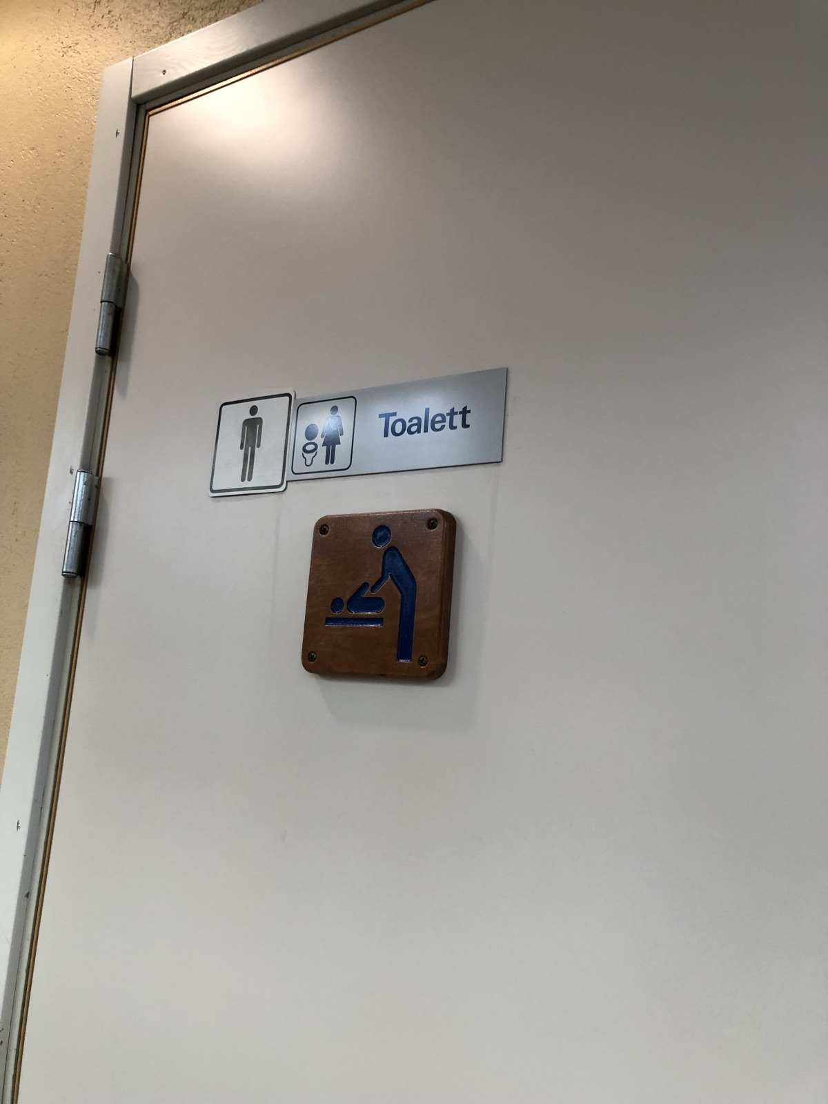 A white toilet door with three signs from different eras. A square sign with a man figure, a silver strip reading Toalett with woman and toilet icons, and below them a worn wooden plaque with a raised blue baby-changing pictogram.