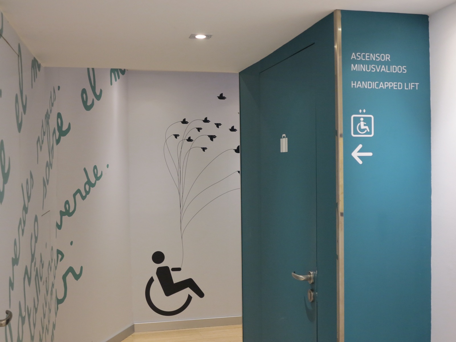 Corridor at a Spanish hotel. On the white wall, a large artwork: a black wheelchair-user figure releasing a flock of birds on long lines. On the teal pillar beside it, a sign reads Ascensor minusválidos, Handicapped lift, with an arrow.