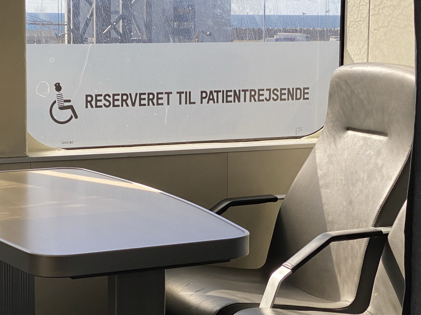Lounge aboard a Danish ferry. Window frosting reads RESERVERET TIL PATIENTREJSENDE, Reserved for patient travellers, beside a wheelchair pictogram drawn with a striped jersey in the line's playful style. Grey leather seat and table.