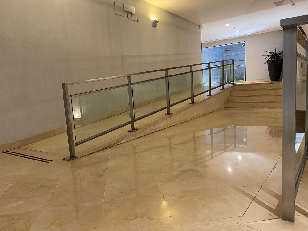 A corridor inside the hotel where a short flight of marble steps is paired with a long ramp with a glass and steel railing.