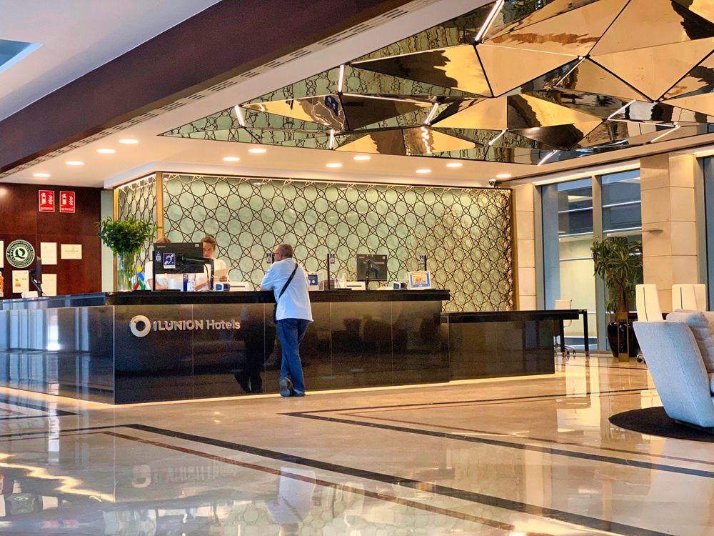 The ILUNION Hotels reception: a dark counter with the hotel name on its front, a guest standing at it and a receptionist behind, under a mirrored faceted ceiling and a patterned metal screen.