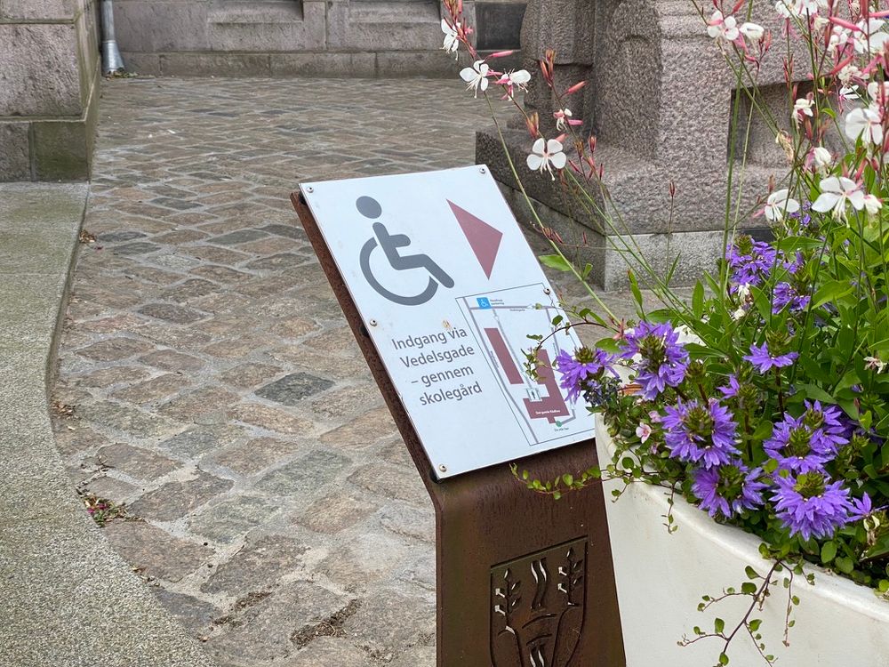 A small sign on a rusted steel post in front of a cobbled ramp and a stone building. A wheelchair symbol, an arrow pointing down and to the right, a small map and the Danish text "Indgang via Vedelsgade – gennem skolegård". A planter with purple flowers stands beside it.