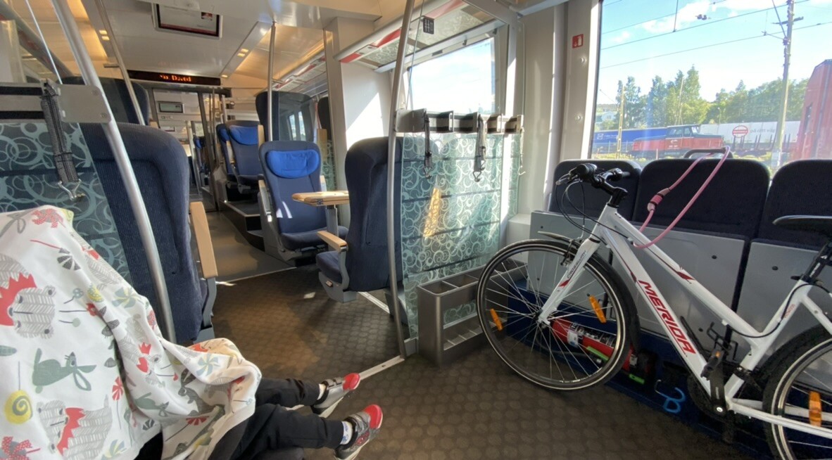 A photograph taken from a wheelchair position inside a train carriage. In the foreground, a cramped space marked with a wheelchair symbol contains a stroller and a bicycle, situated next to a large toilet wall. In the background, through a narrow passage, the main passenger area is visible with comfortable face-to-face seating, tables, and power outlets, highlighting the physical segregation between "typical" passengers and those on wheels.