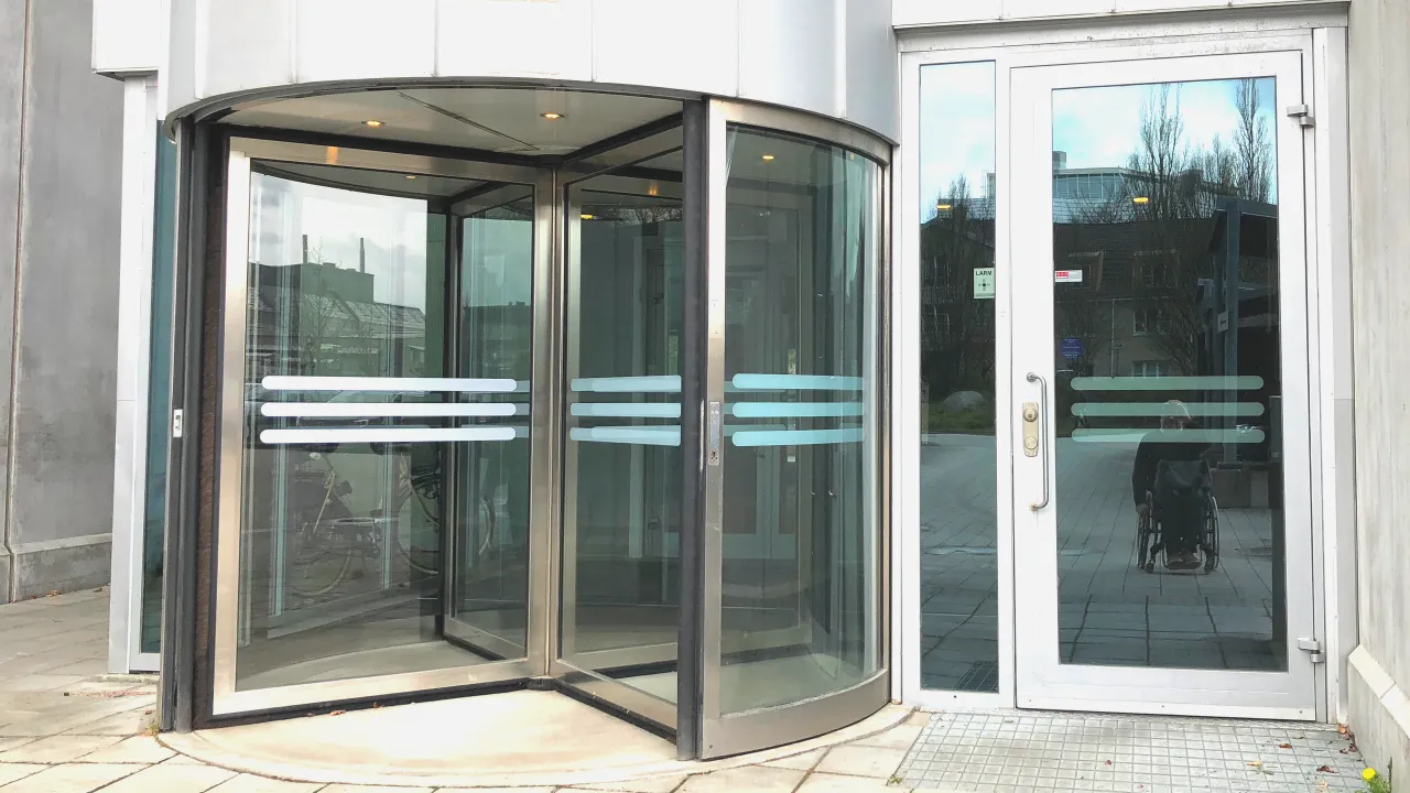 An entrance with a revolving door to the left and a straight door to the right. There is no wheelchair sign, but my wheelchair is reflected back at me in the glass of the straight door.