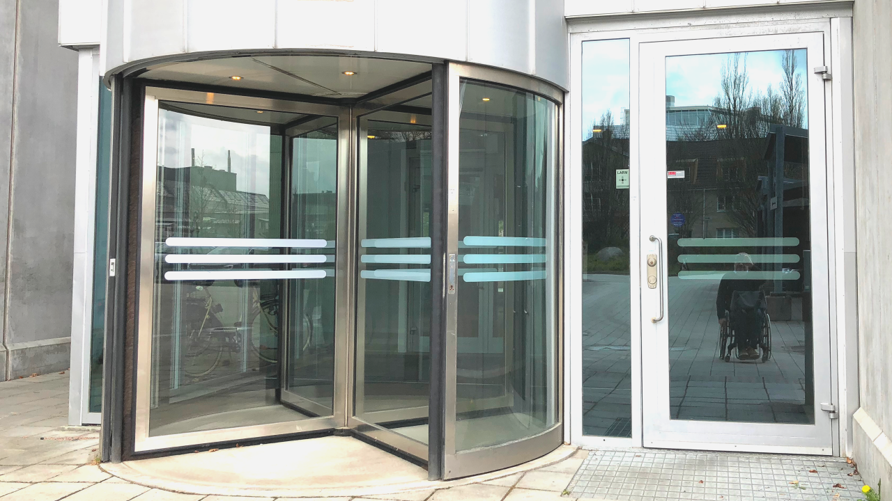 An entrance with a revolving door to the left and a straight door to the right. There is no wheelchair sign, but my wheelchair is reflected back at me in the glass of the straight door.