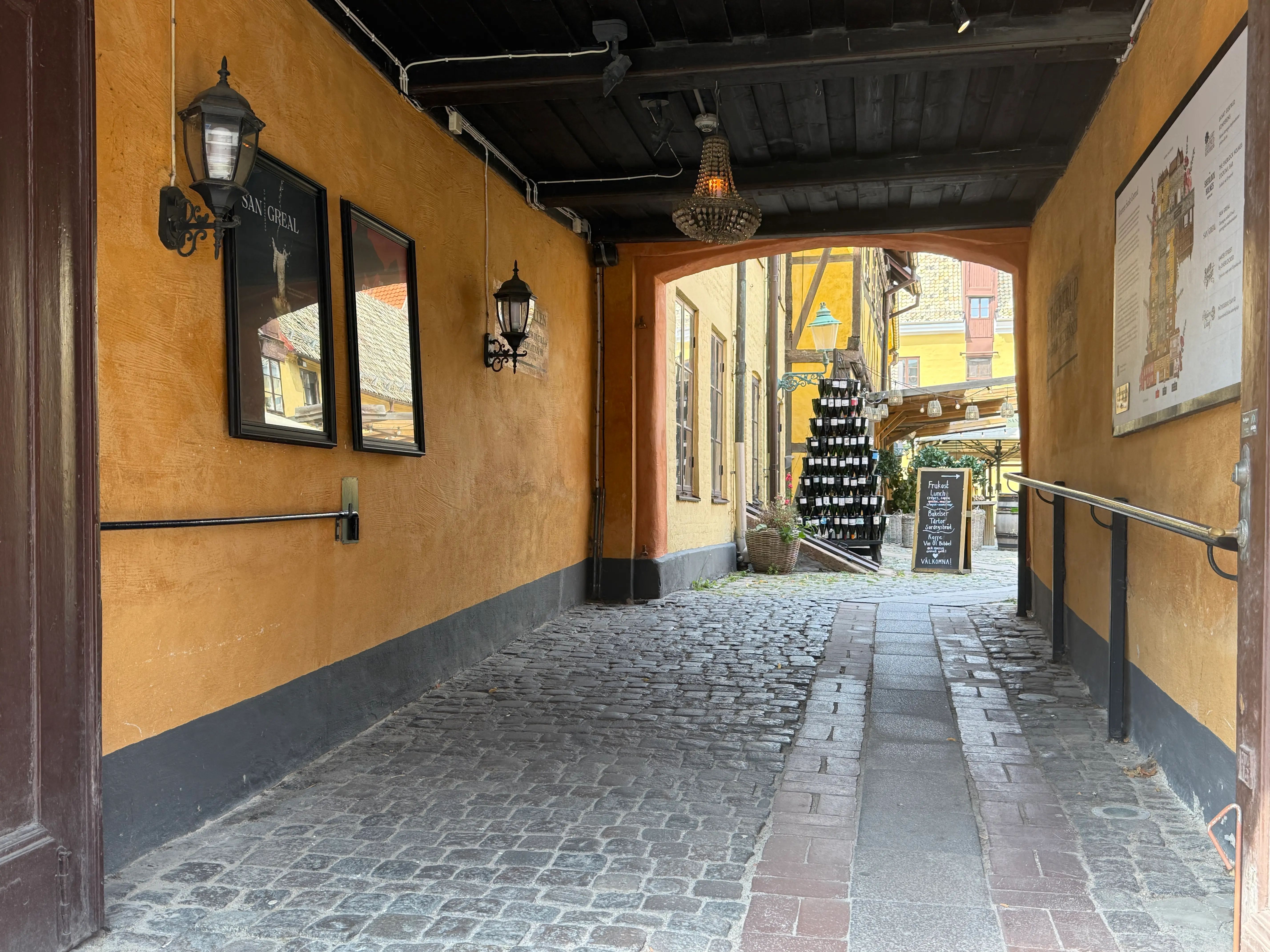 I am entering a covered passage into a restaurant courtyard in a heritage quarter in Malmö. A band of smooth stones is laid through the rough cobblestones, with a metal handrail along the ochre wall.