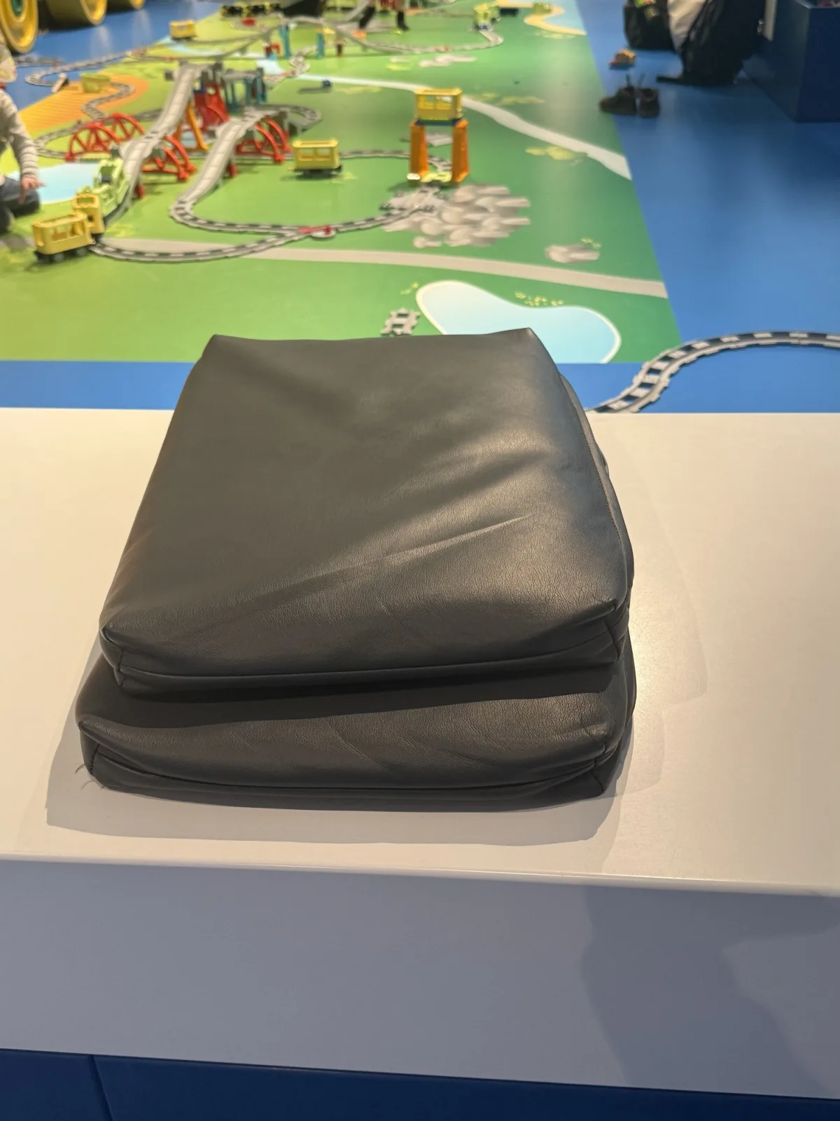Two black cushions lie stacked on the white border around a large Duplo train landscape. Behind them are tracks, bridges and small trains on a green play mat.