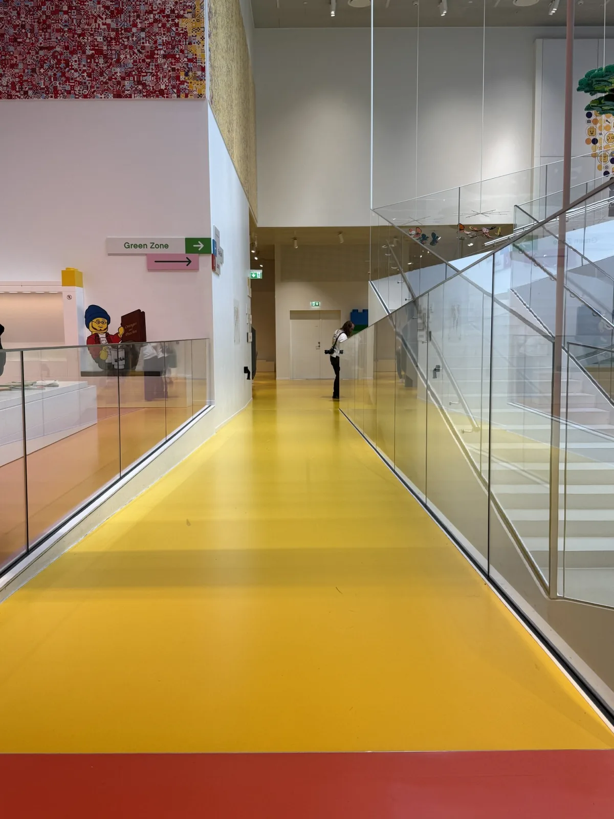 A wide yellow floor ramps up toward the viewer inside a white building, with glass balustrades on both sides. Behind the glass on the right, a white staircase runs beside the ramp and rises the other way. A sign on the wall reads Green Zone, with an arrow. A red floor meets the top of the ramp in the foreground, and a person with a camera stands at the bottom, at the far end.