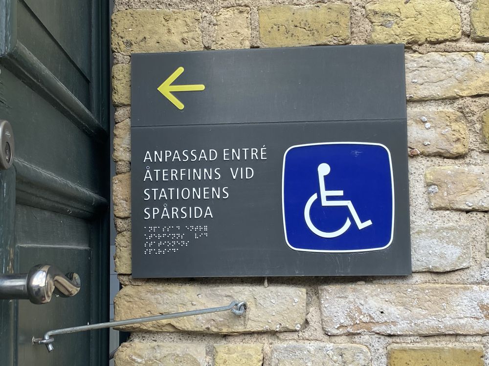 A dark sign on a yellow brick wall beside a green door. A yellow arrow points left. Below it, the Swedish text "Anpassad entré återfinns vid stationens spårsida" with braille, and a blue wheelchair symbol.